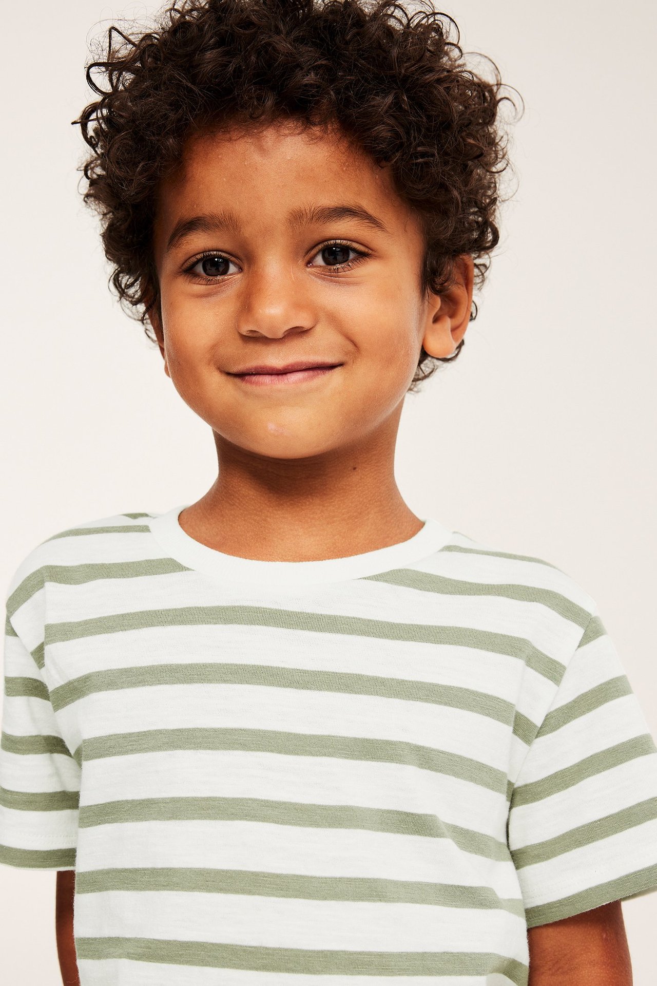 Närbild framifrån på ett leende barn i en vit och mörkgrön randig t-shirt i bomull.