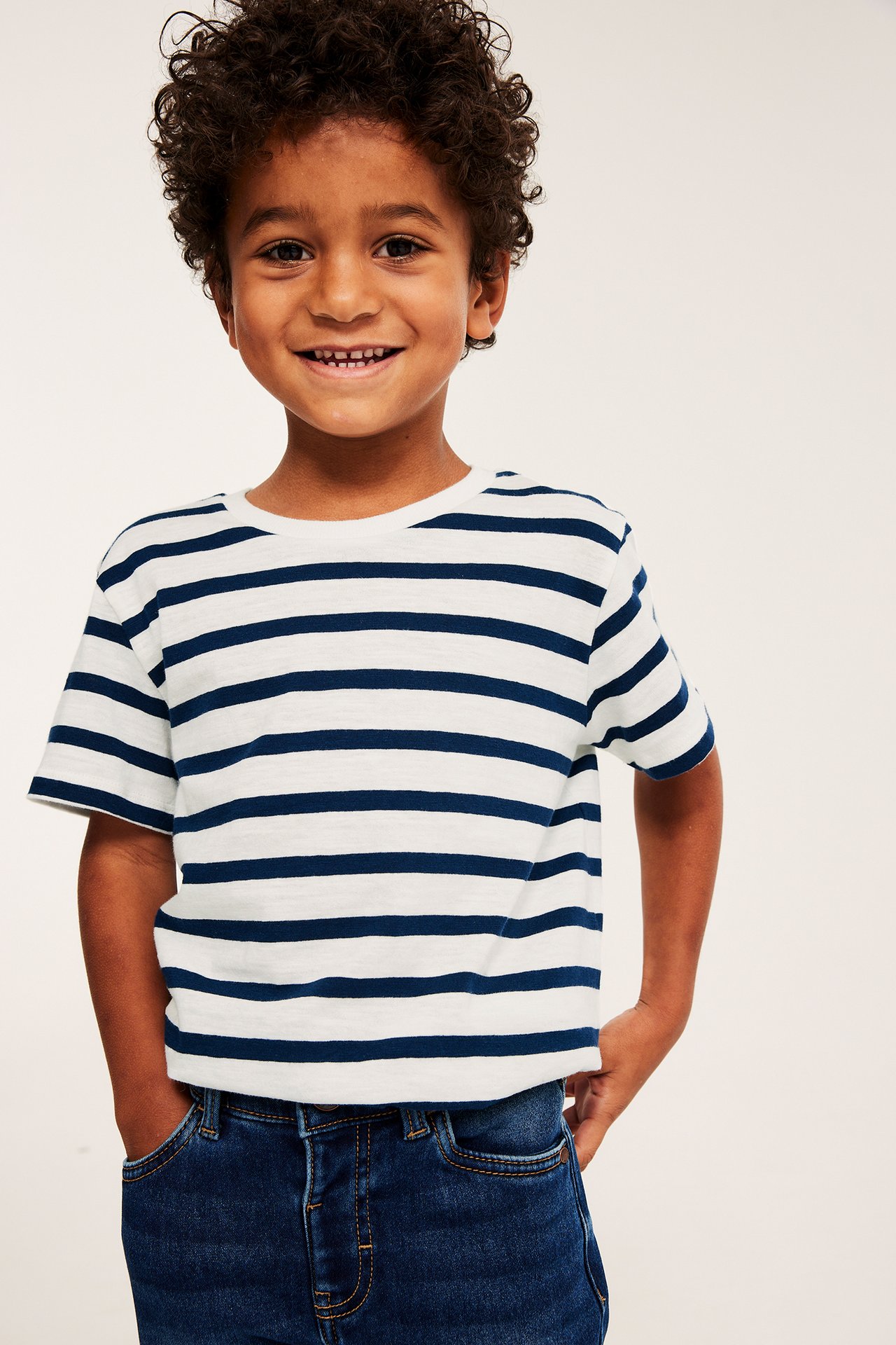 Forfra: et smilende barn i en hvit og mørkeblå stripete bomulls-t-skjorte og mørkeblå jeans.