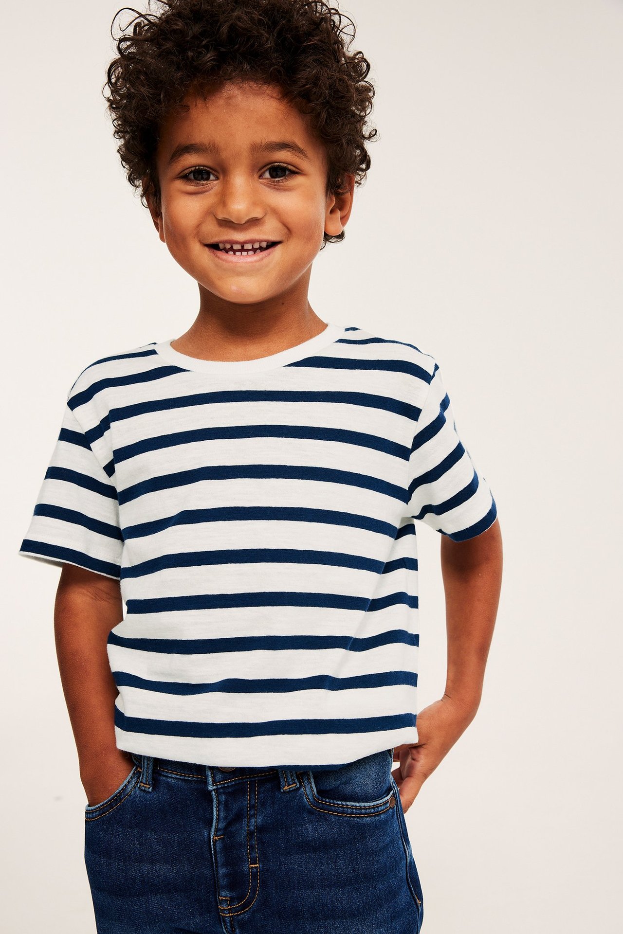 Framifrån: leende barn i vit och mörkblå randig bomulls-t-shirt och mörkblå jeans.