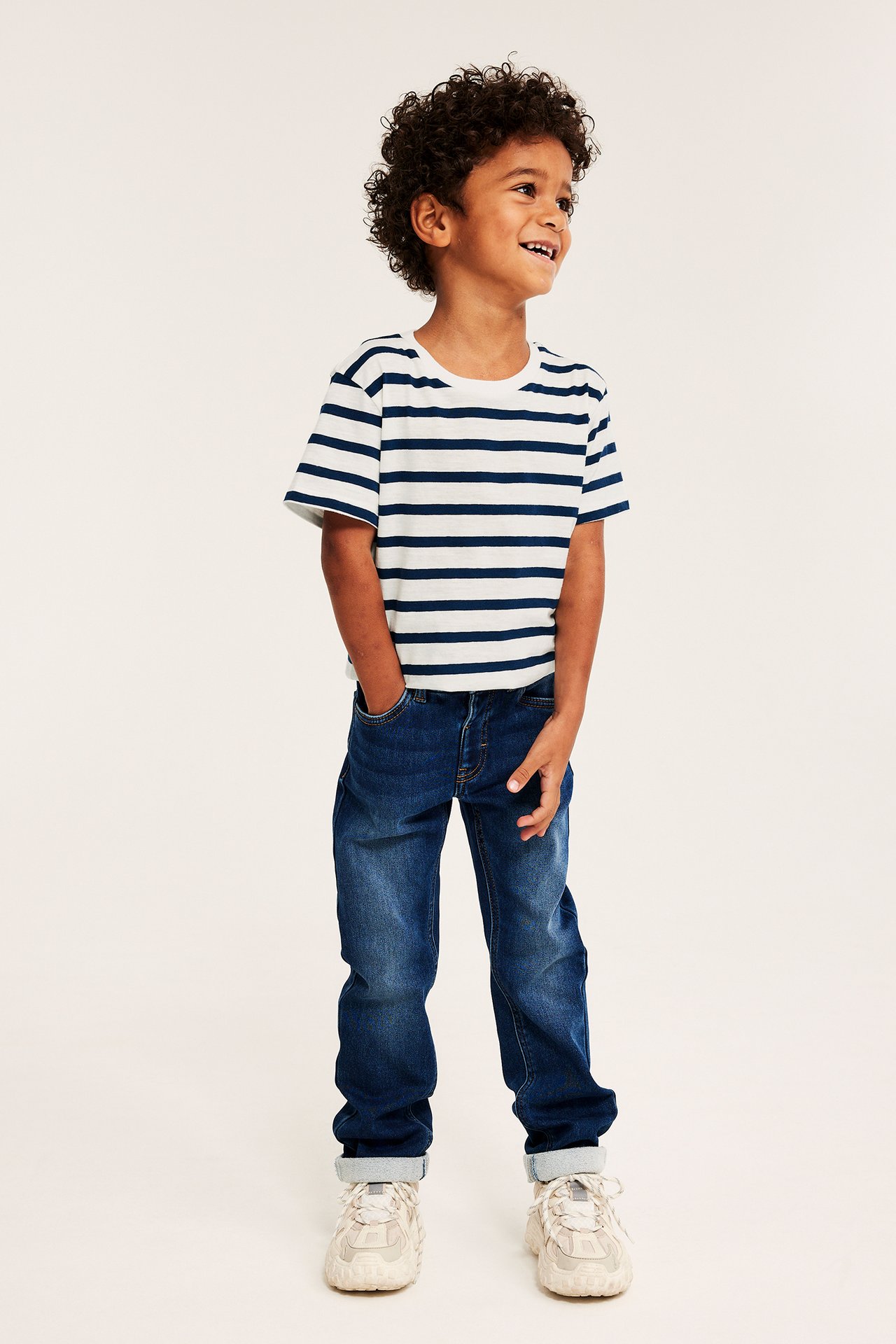 Dziecko w biało-granatowym, pasiastym T-shircie, granatowych jeansach i beżowych trampkach.