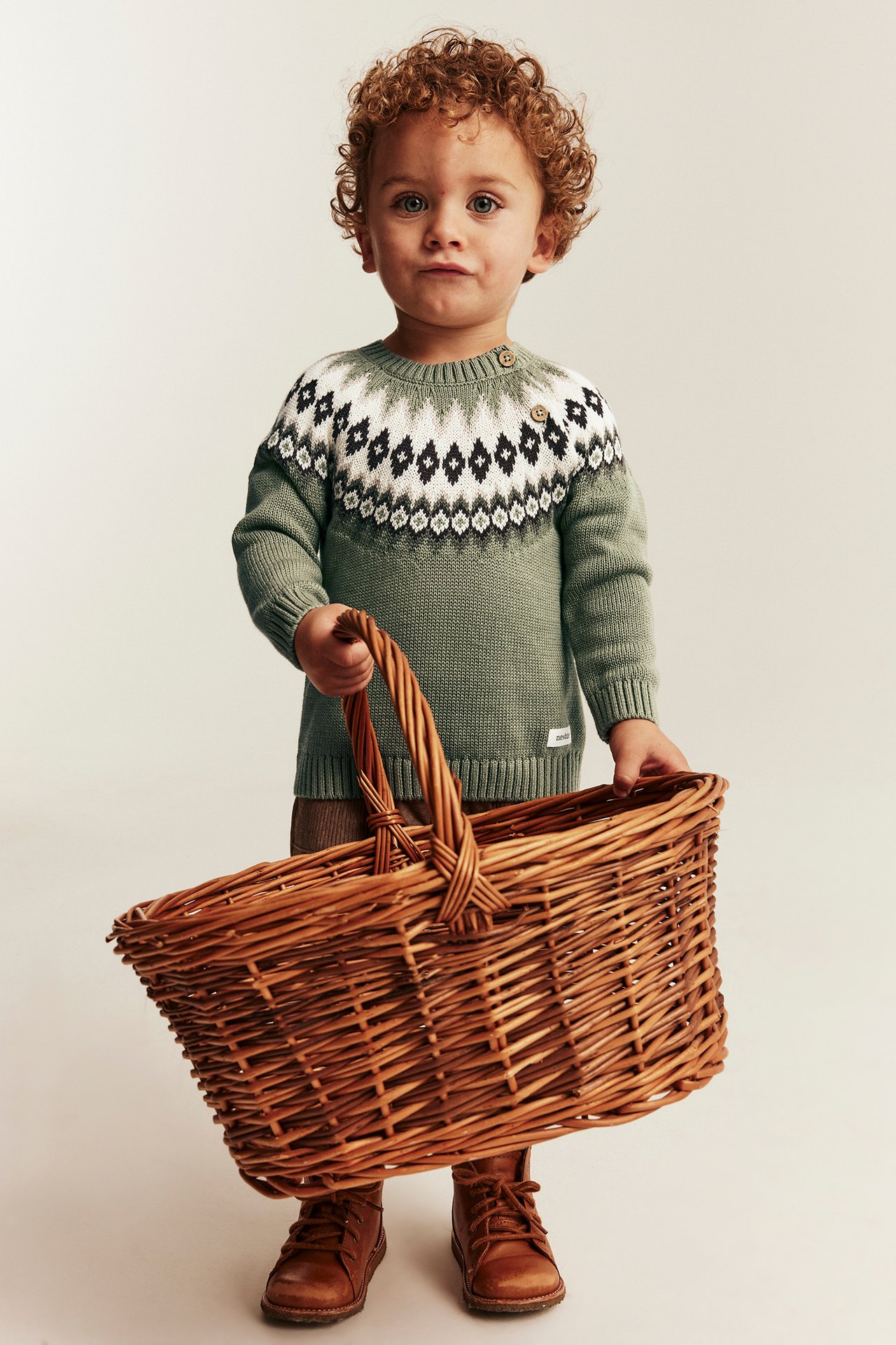 Barn i grønn fair isle-genser med svart og hvitt, brune bukser, støvler, holder en kurv.