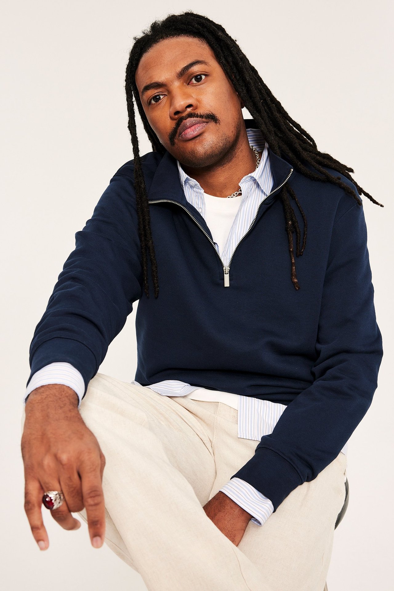 Man i mörkblå half-zip sweatshirt över randig skjorta, vit t-shirt, beiga byxor, silverkedja och röd ring.