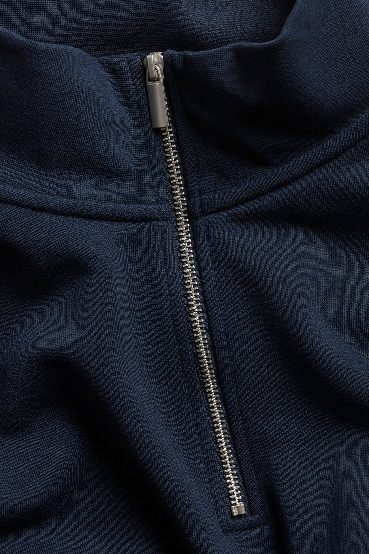 Nærbilde av en mørkeblå bomullssweatshirt med halv glidelås, høy krage og sølvfarget glidelås.