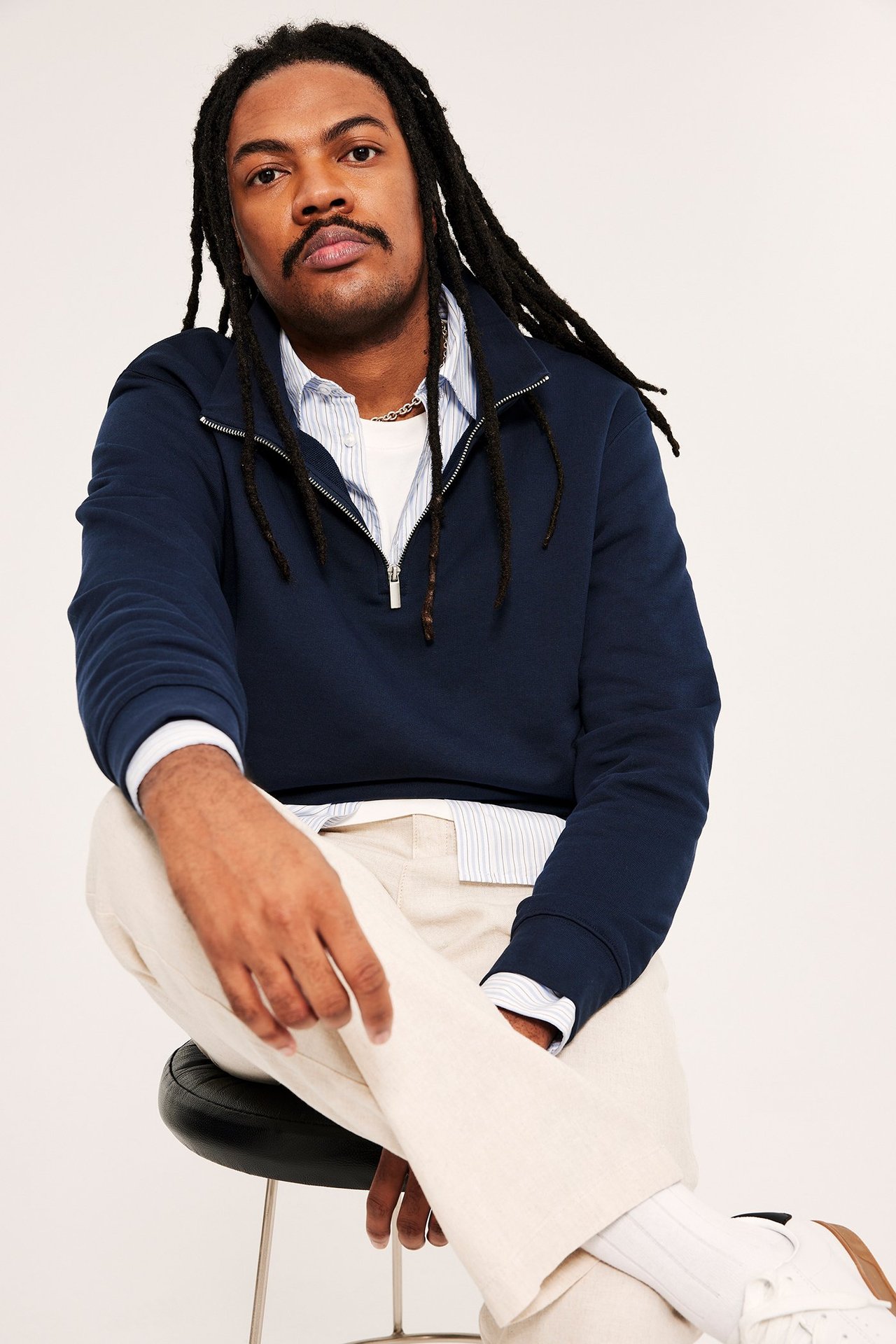 Man bär mörkblå half-zip sweatshirt över randig skjorta, vit t-shirt, beiga byxor och vita sneakers.