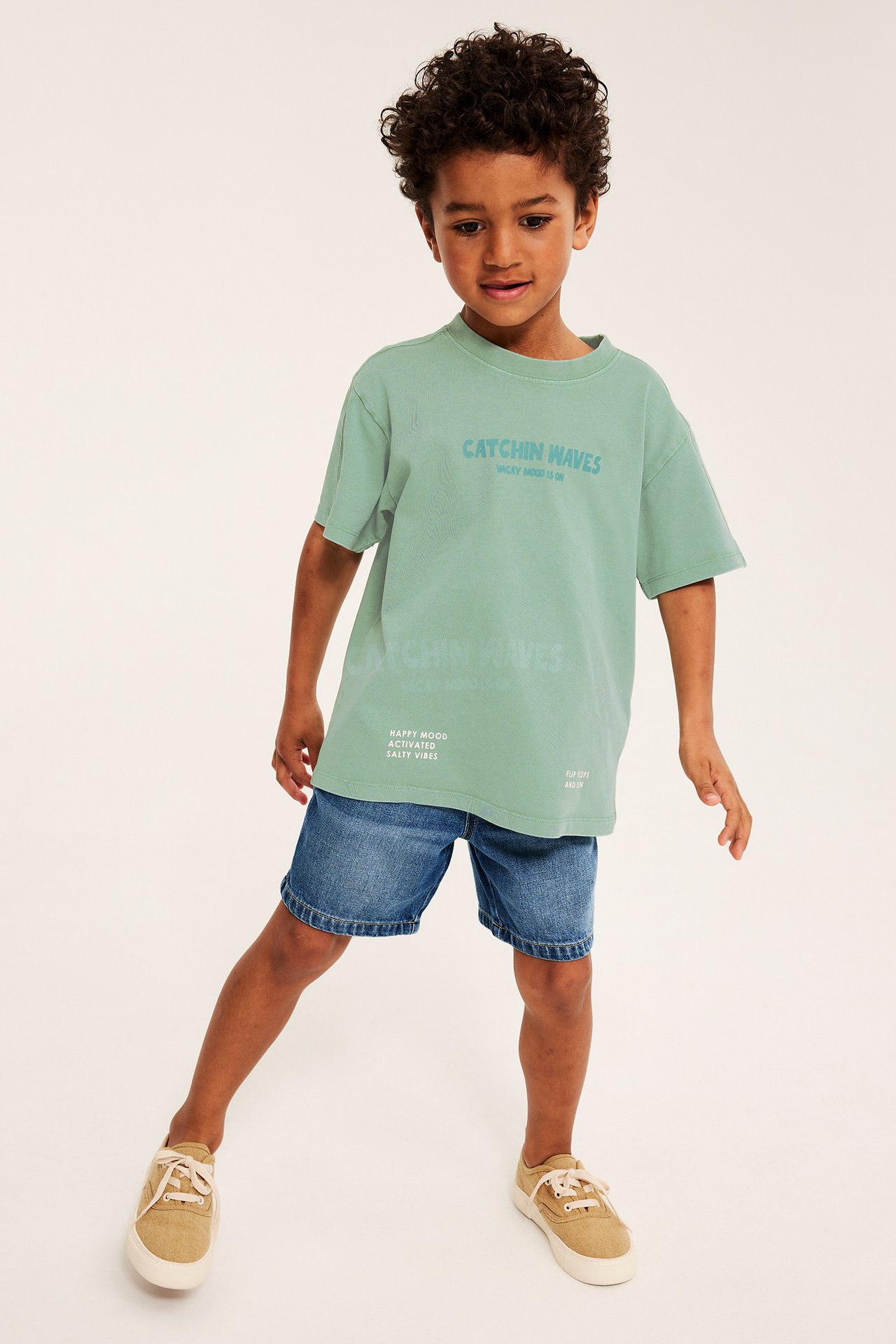 Frontvy på ett barn i en grön, oversized bomulls-t-shirt med texttryck, blå denimshorts och beiga sneakers.