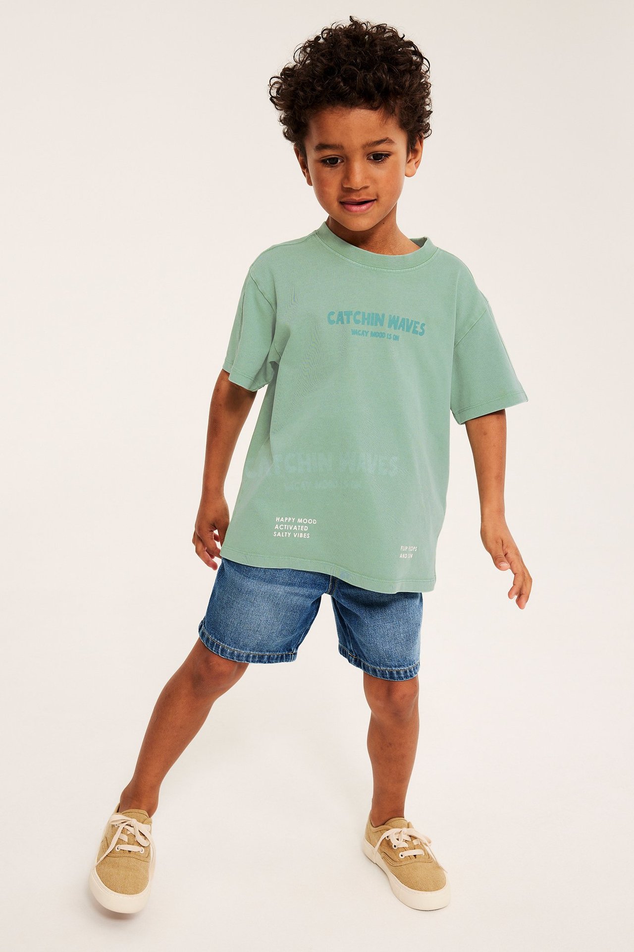 Edestä näkymä lapsesta vihreässä ylisuuressa T-paidassa tekstillä, sinisissä farkkushortseissa ja beigessä tennareissa.