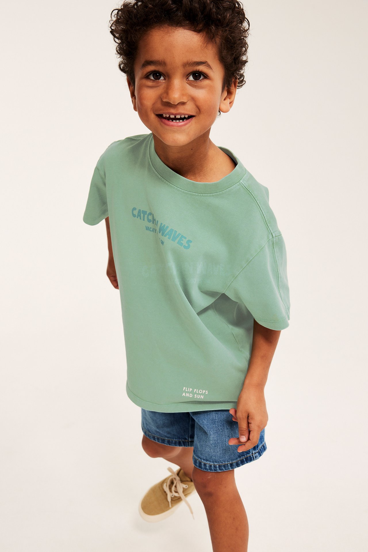 Framifrån, ett barn i en oversize grön bomulls-t-shirt med tryck, blå denimshorts och beiga skor.