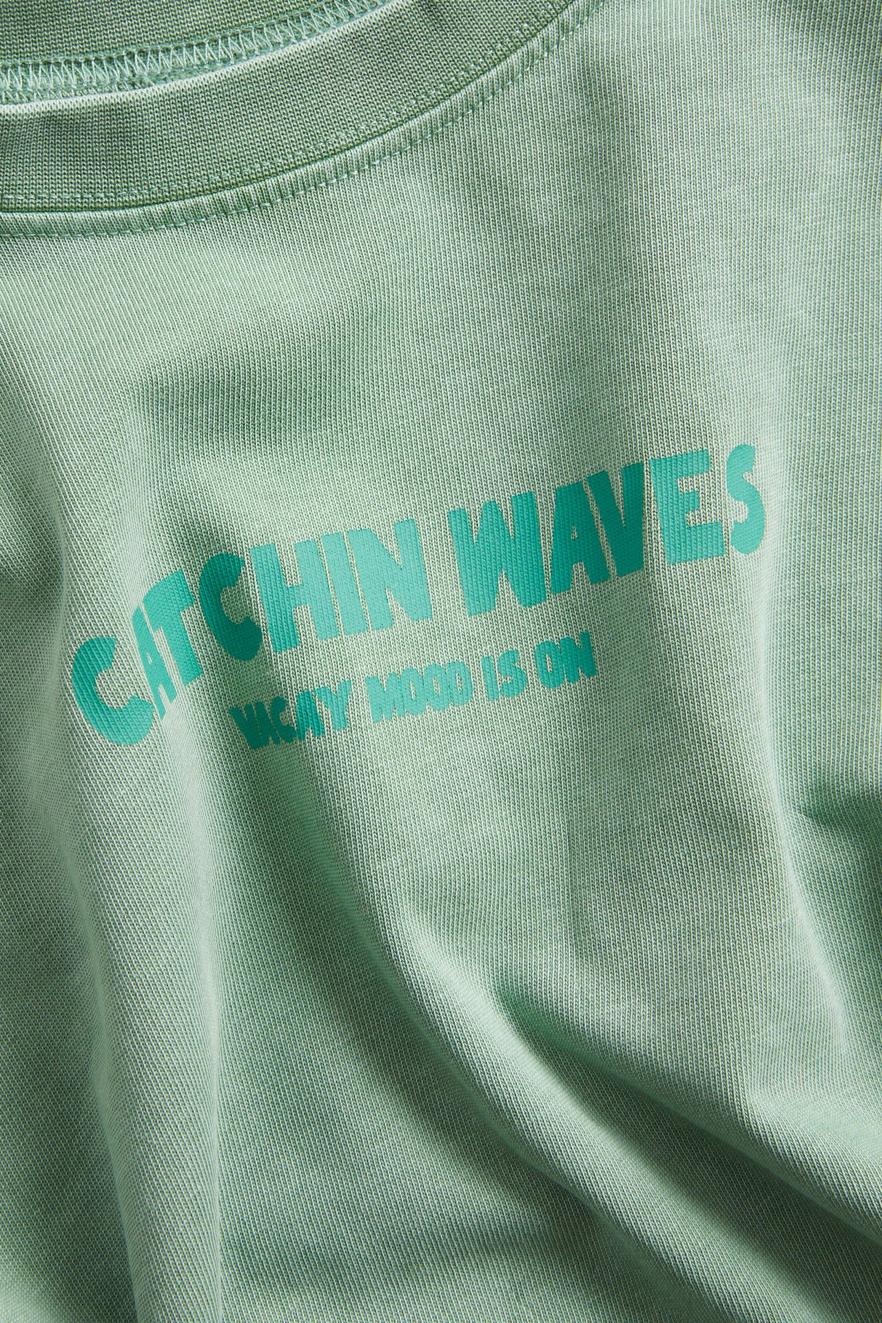 Lähikuva edestä vihreästä oversize puuvilla-t-paidasta, jossa teksti 'CATCHIN WAVES'.