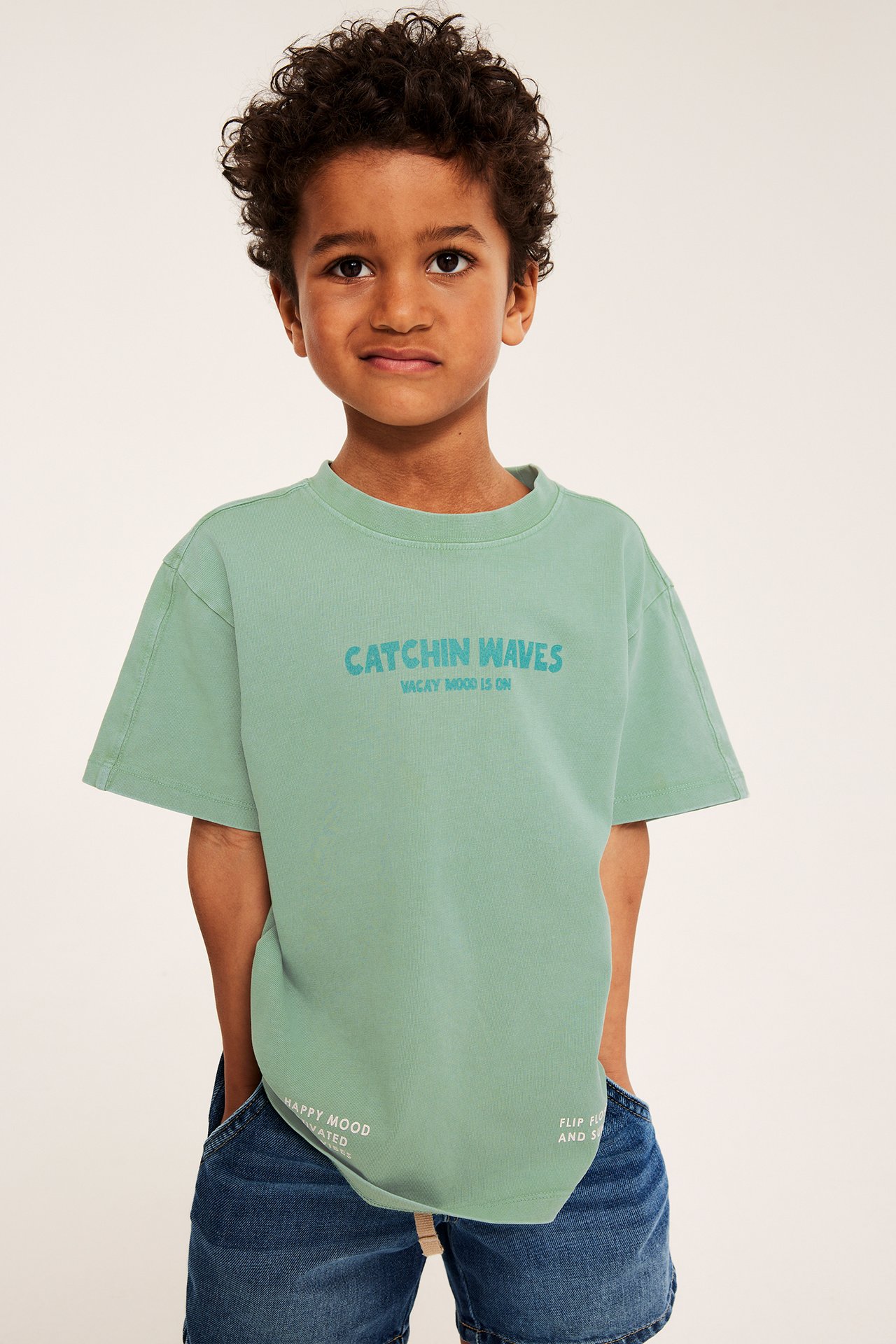 Frontvy av ett barn som bär en grön oversize t-shirt i bomull med blått tryck och blå denimshorts.