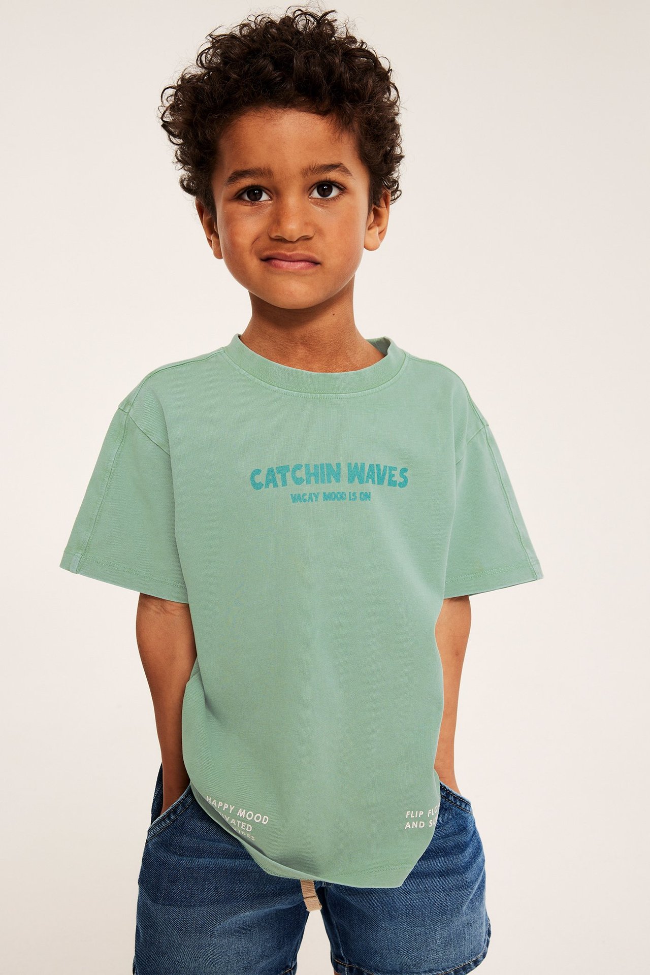 Widok z przodu dziecka w zielonej bawełnianej koszulce oversize z niebieskim nadrukiem i jeansowych szortach.