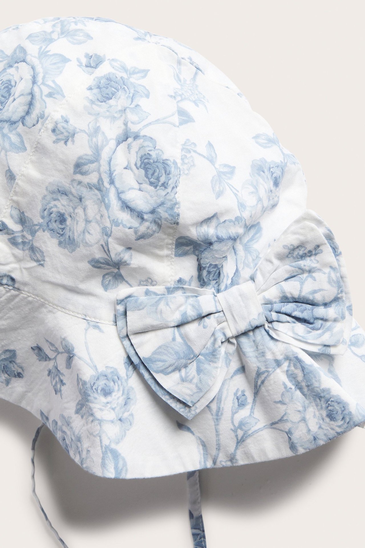 Nærbilde forfra av en offwhite babysolhatt med blått blomstermønster, med stor sløyfe og knytebånd.