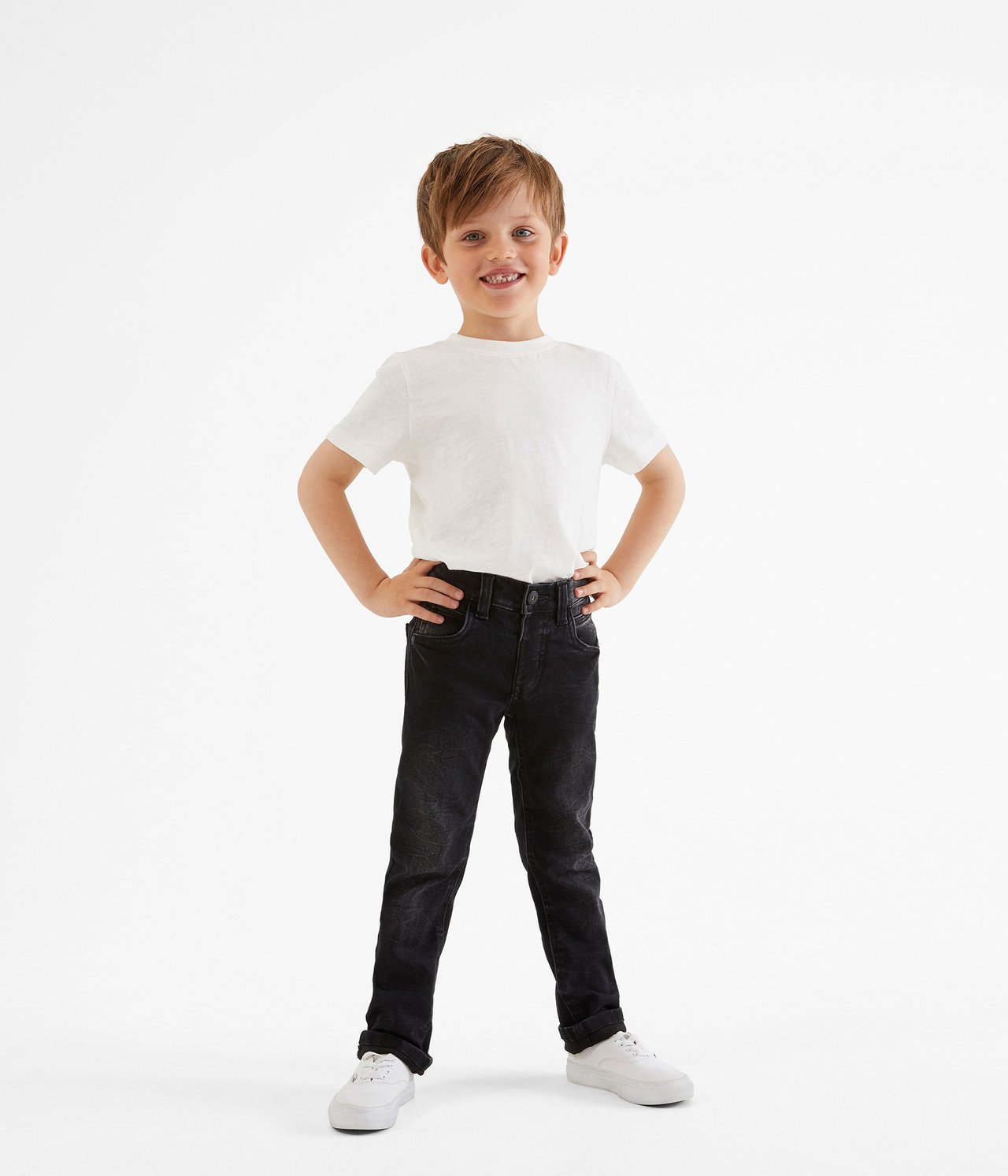 Framifrån: ett barn i svarta slim-fit joggerjeans, en vit t-shirt och vita sneakers.