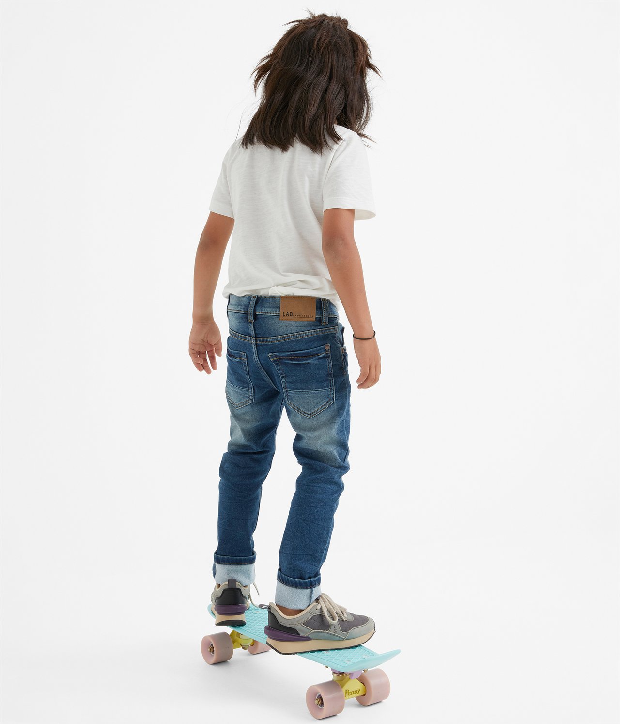 Takaapäin: Lapsi slim fit denim joggerfarkuissa, valkoisessa t-paidassa, harmaissa lenkkareissa, rullalaudalla.