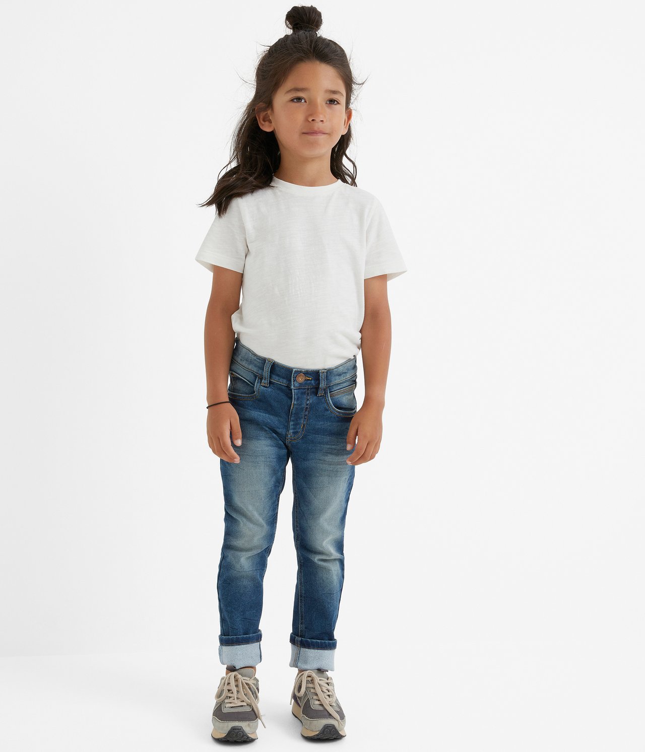 Edestäpäin: lapsi yllään siniset slim-fit joggerfarkut käärityillä lahkeilla, valkoinen t-paita ja harmaat lenkkarit.