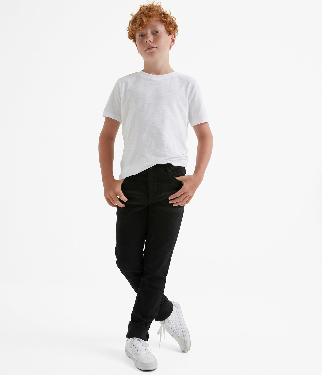 Widok z przodu dziecka w czarnych dżinsach jogger slim-fit, białym T-shircie i białych trampkach.