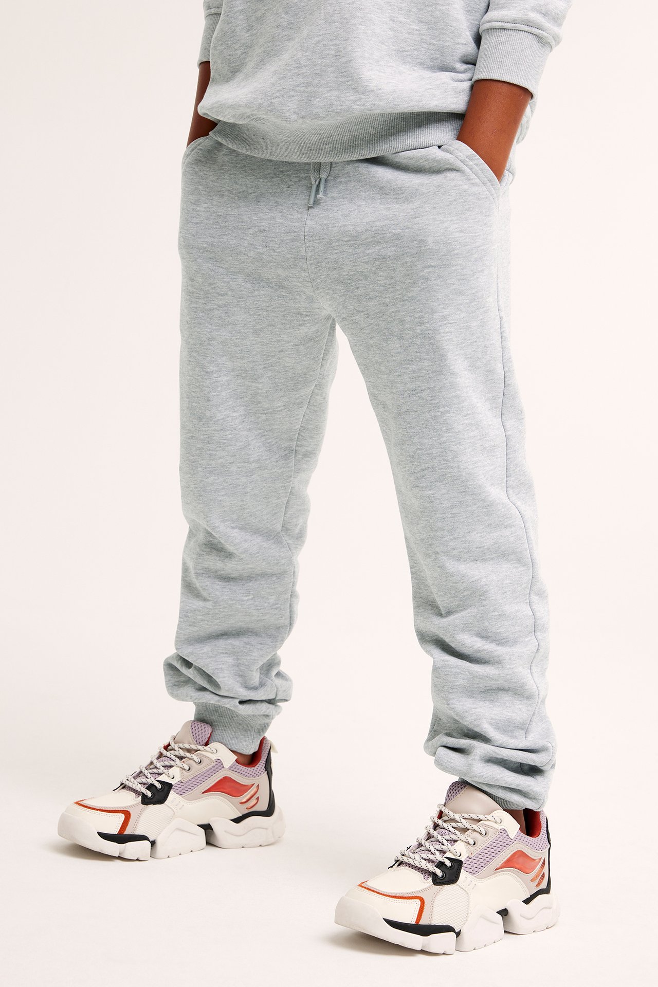 Framifrån: Ett barn bär gråmelerade mjukisbyxor med dragsko, fickor, muddar och chunky sneakers.