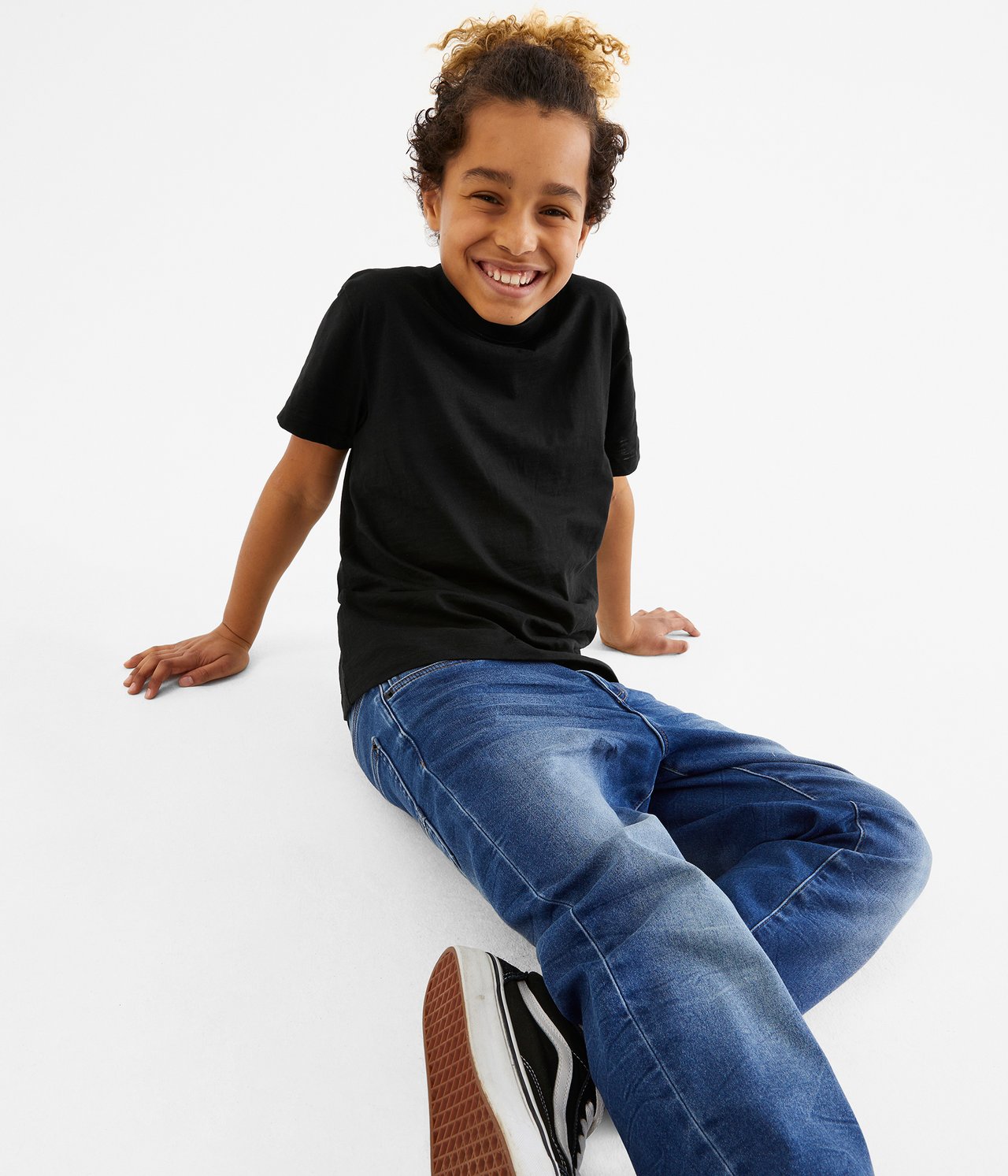 Framifrån, leende barn i svart bomulls-T-shirt, blå jeans och svartvita sneakers.