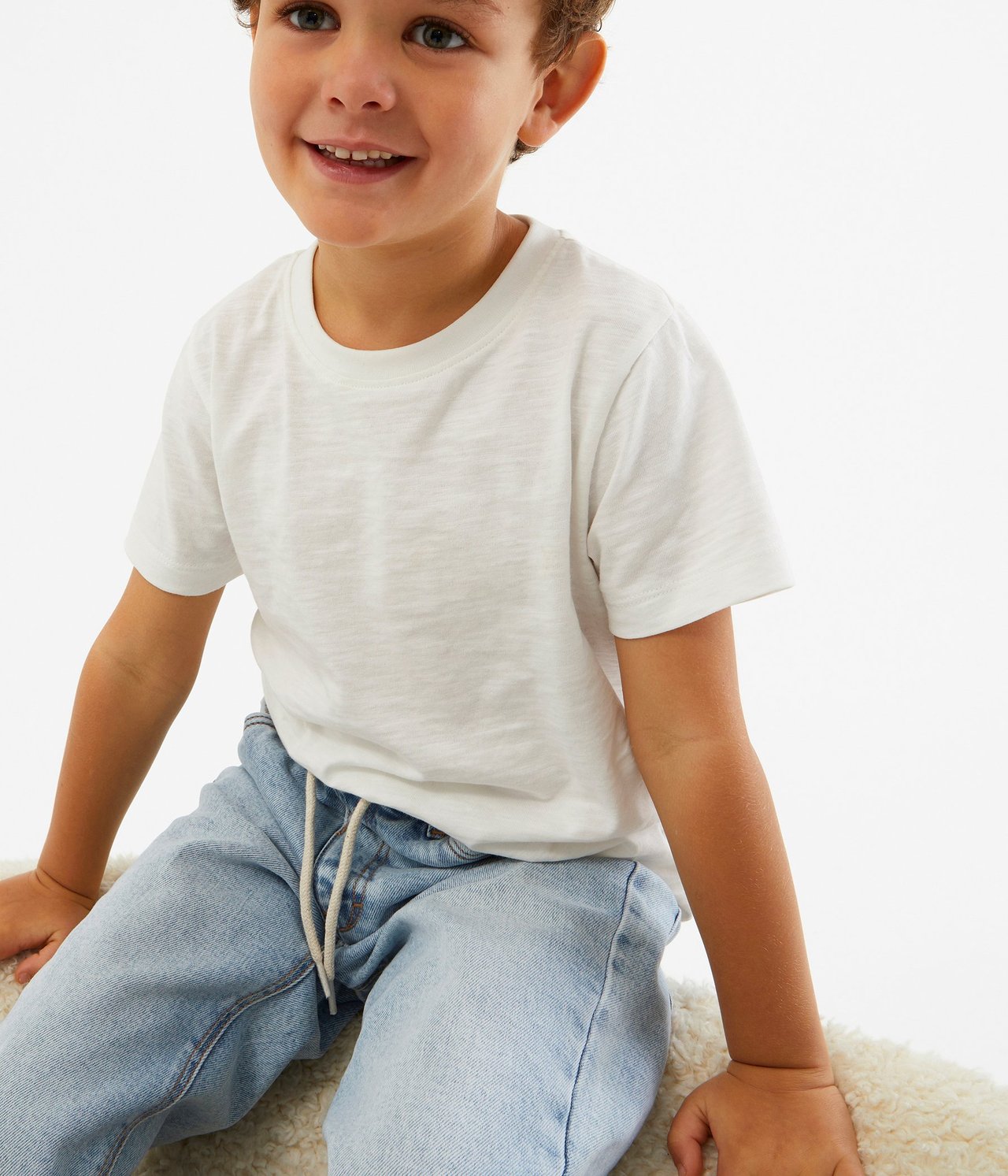 Edestä, hymyilevä lapsi yllään luonnonvalkoinen puuvillainen t-paita ja vaaleansiniset farkut.