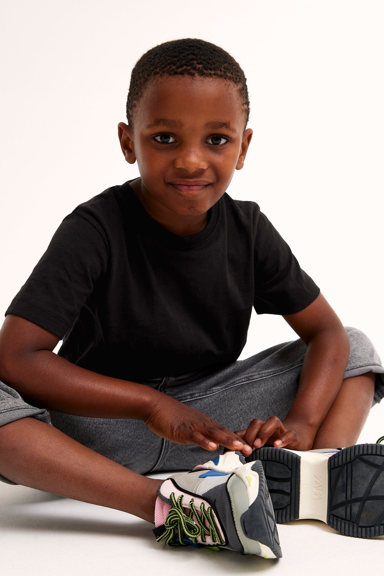 Framifrån: ett barn i svart bomulls-t-shirt, grå byxor och flerfärgade sneakers med tjock sula.