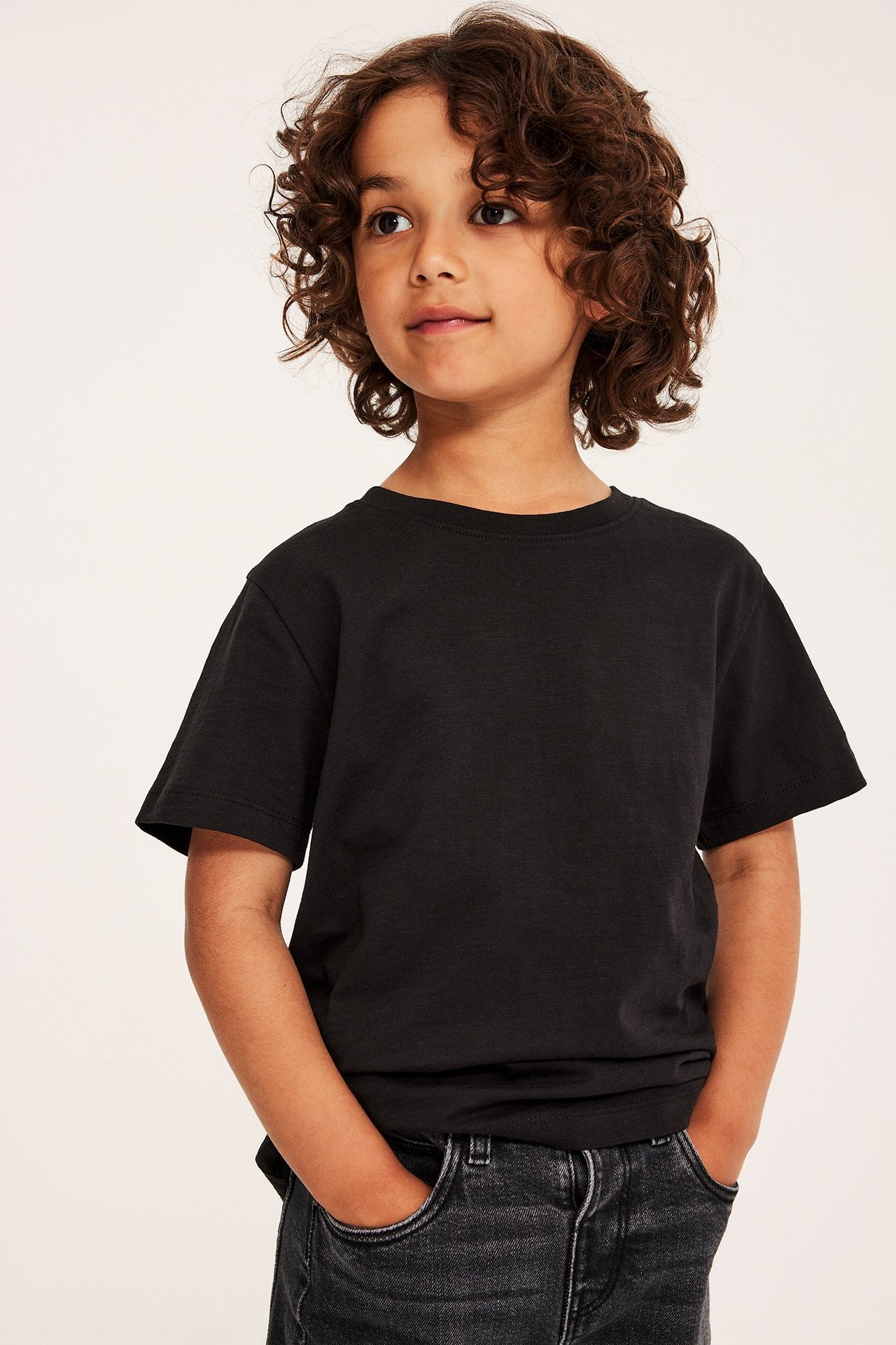 Widok z przodu dziecka w czarnym bawełnianym T-shircie z okrągłym dekoltem i ciemnoszarych dżinsach.