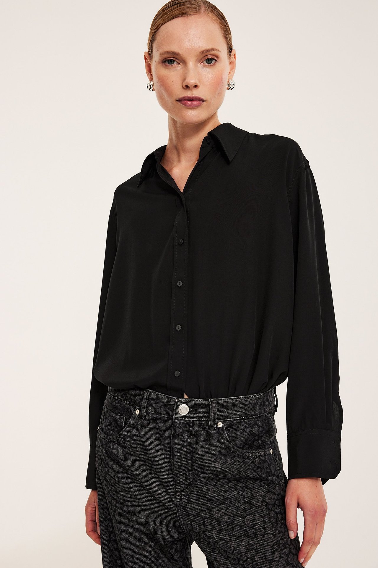 Widok z przodu: Kobieta w czarnej koszuli oversize, ciemnych jeansach w panterkę i srebrnych kolczykach.
