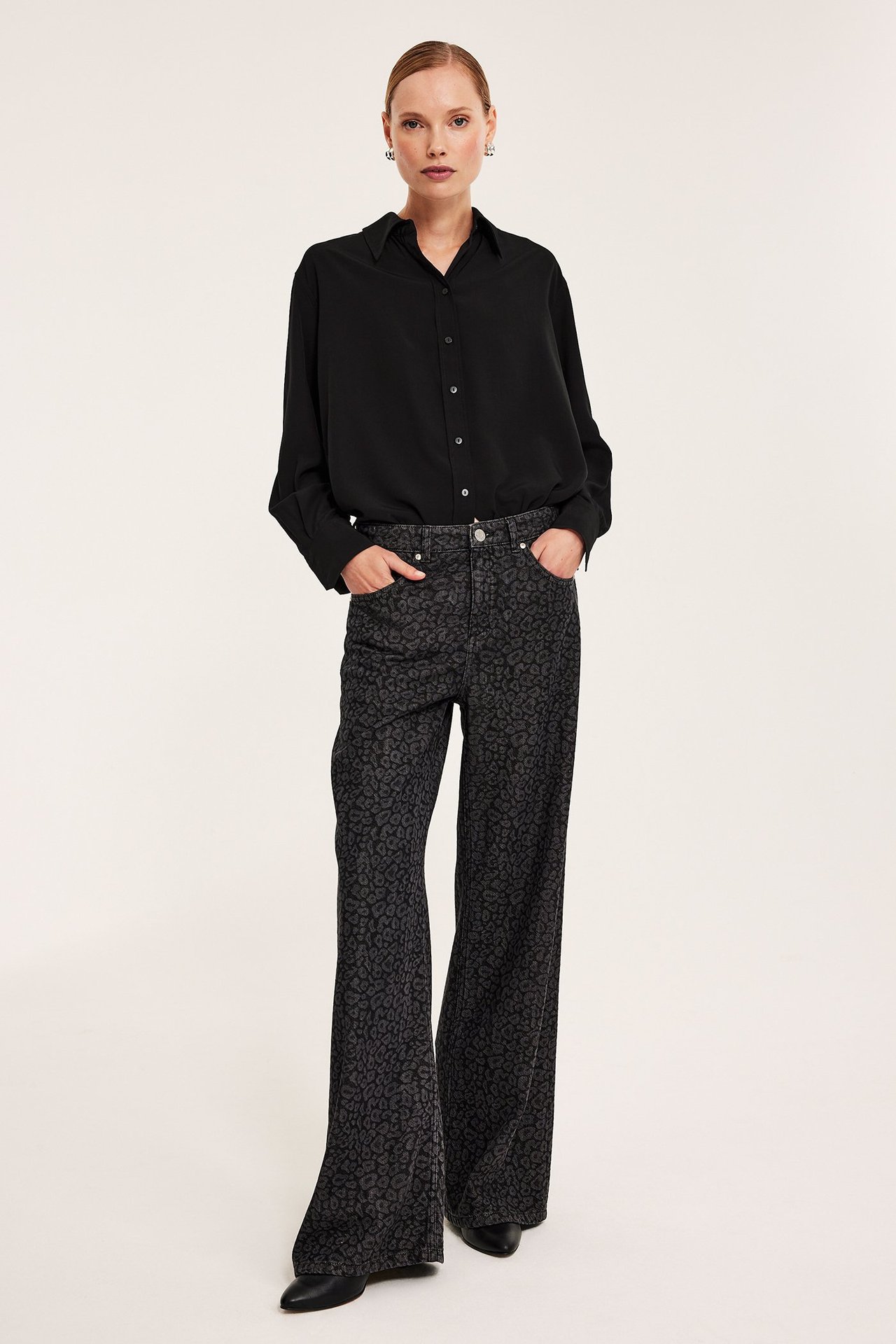 Pełny widok z przodu kobiety w czarnej koszuli oversize z wiskozy, ciemnoszarych spodniach w zwierzęcy wzór i czarnych butach.