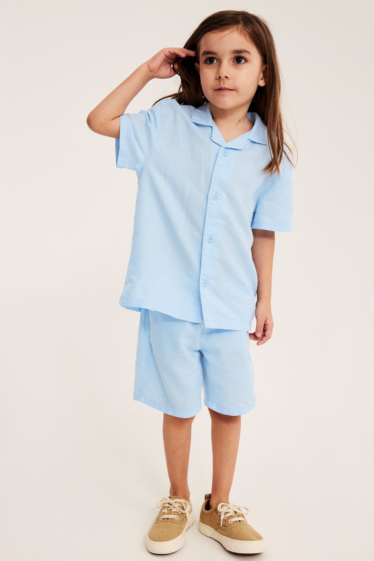 Framifrån: kid bär ljusblå kortärmad linneskjorta, matchande shorts och beige canvasskor.