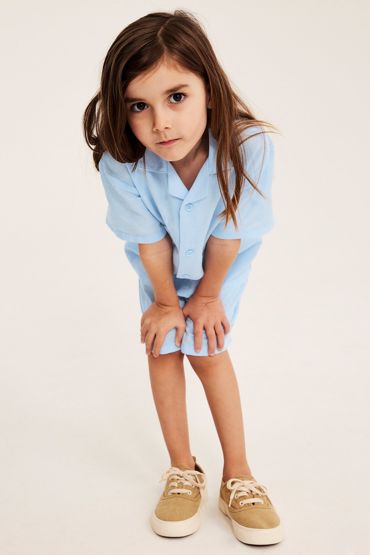 Frontvy av ett barn i ljusblå kortärmad resortsjorta, matchande shorts och beiga canvassneakers.
