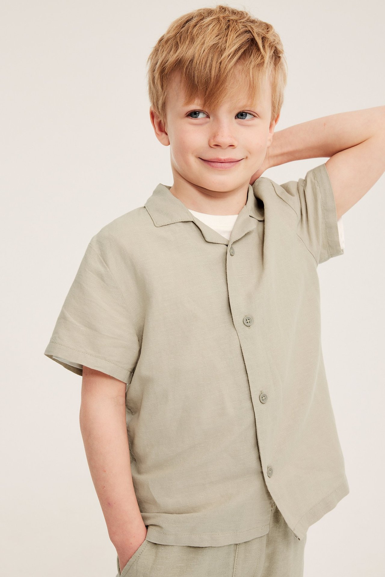 Från framsidan, ett barn bär en beige kortärmad linneskjorta över vit t-shirt och matchande shorts.