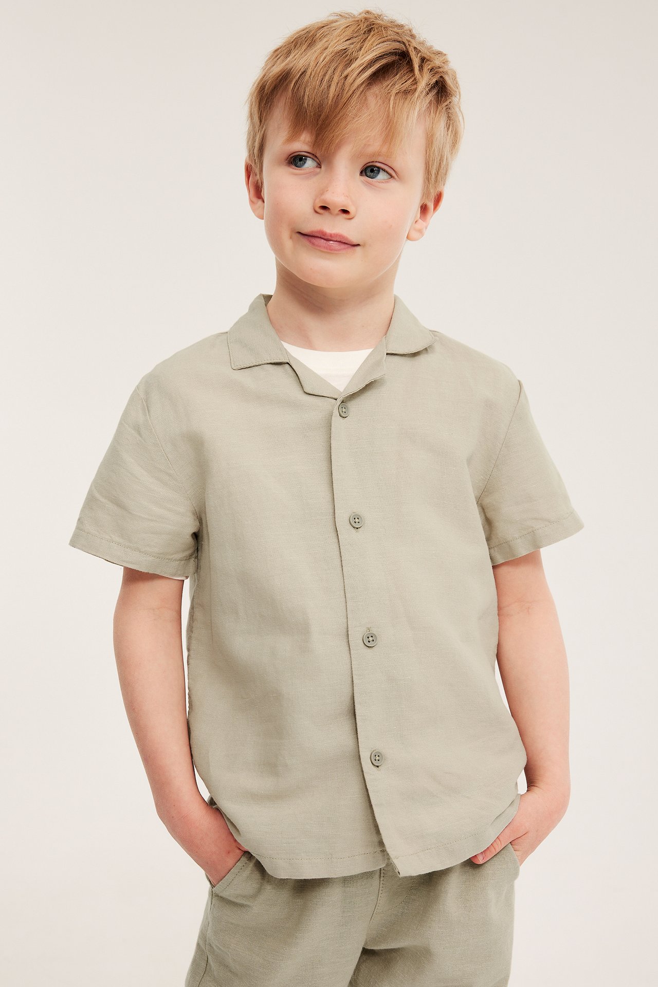 Widok z przodu: Chłopiec w beżowej lnianej koszuli z krótkim rękawem, białym T-shircie i pasujących beżowych szortach.