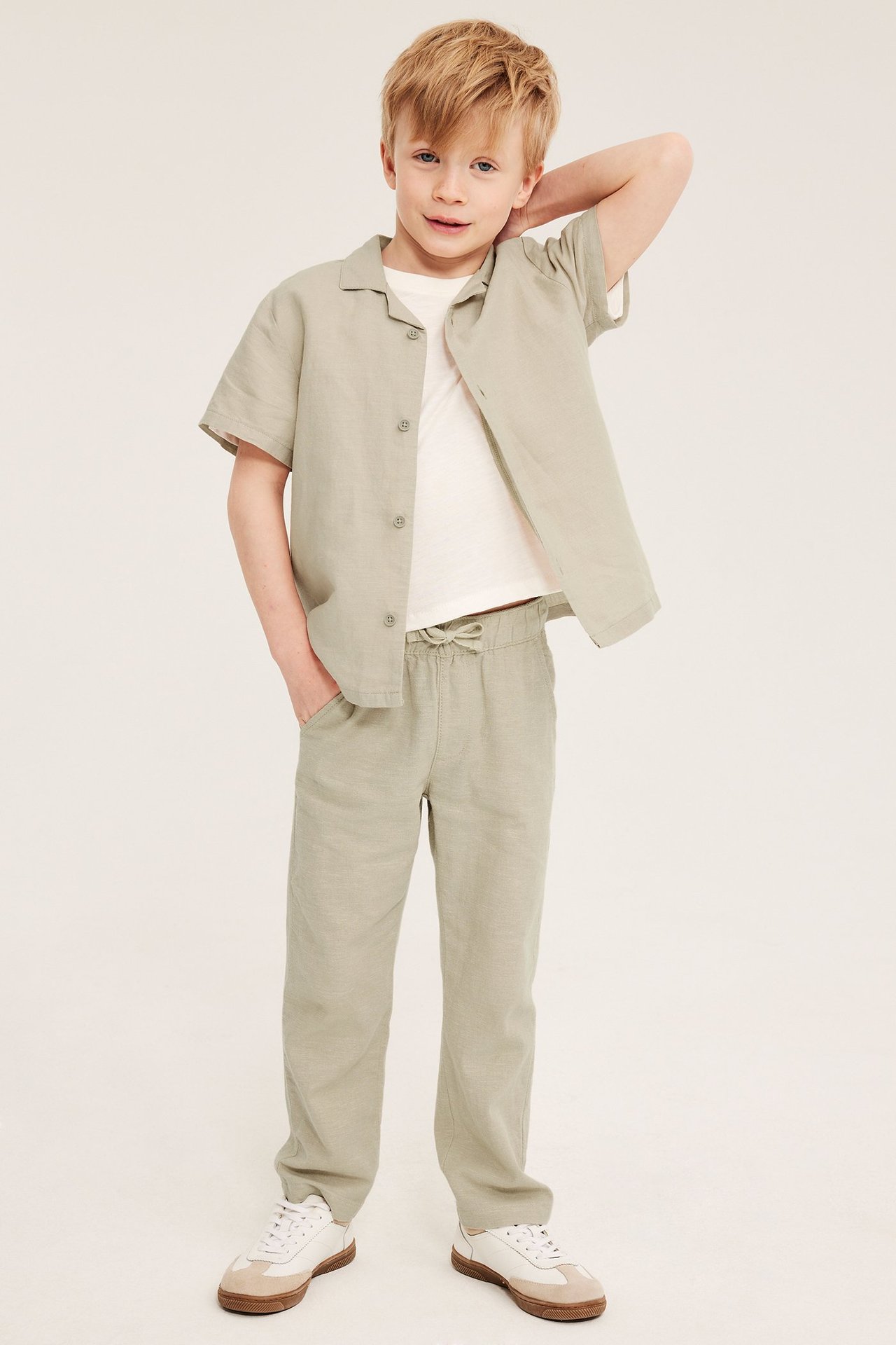Widok z przodu: dziecko w rozpiętej beżowej lnianej koszuli, białym T-shircie, pasujących spodniach i sneakersach.