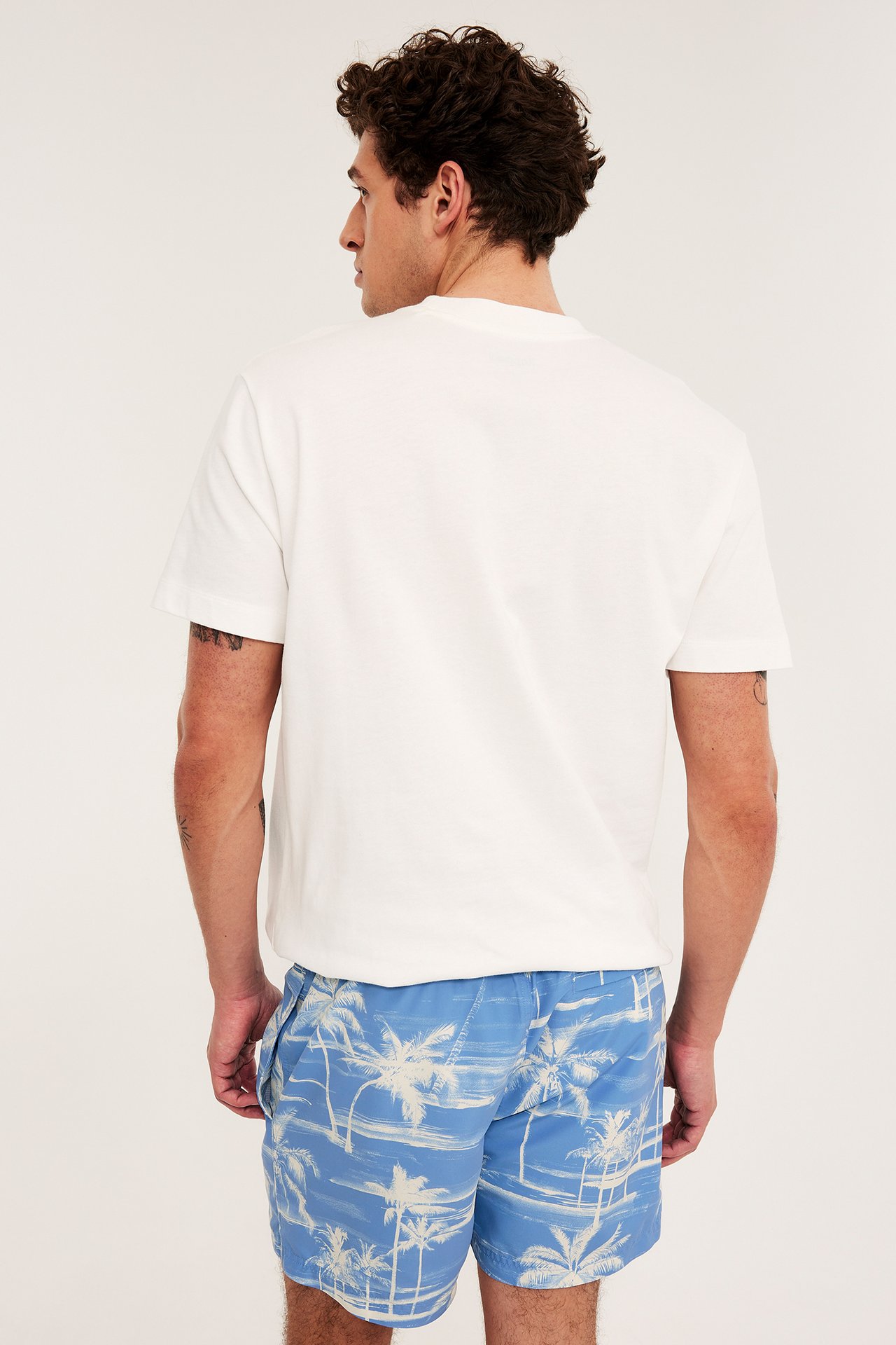 Man i ryggvy bär ljusblå palm-mönstrade badshorts och en enkel vit t-shirt.