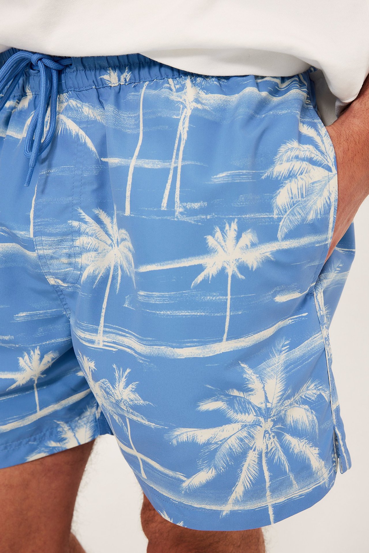Lähikuva miehestä yllään vaaleansiniset uimahousut valkoisella palmupuukuvioinnilla ja valkoinen t-paita.
