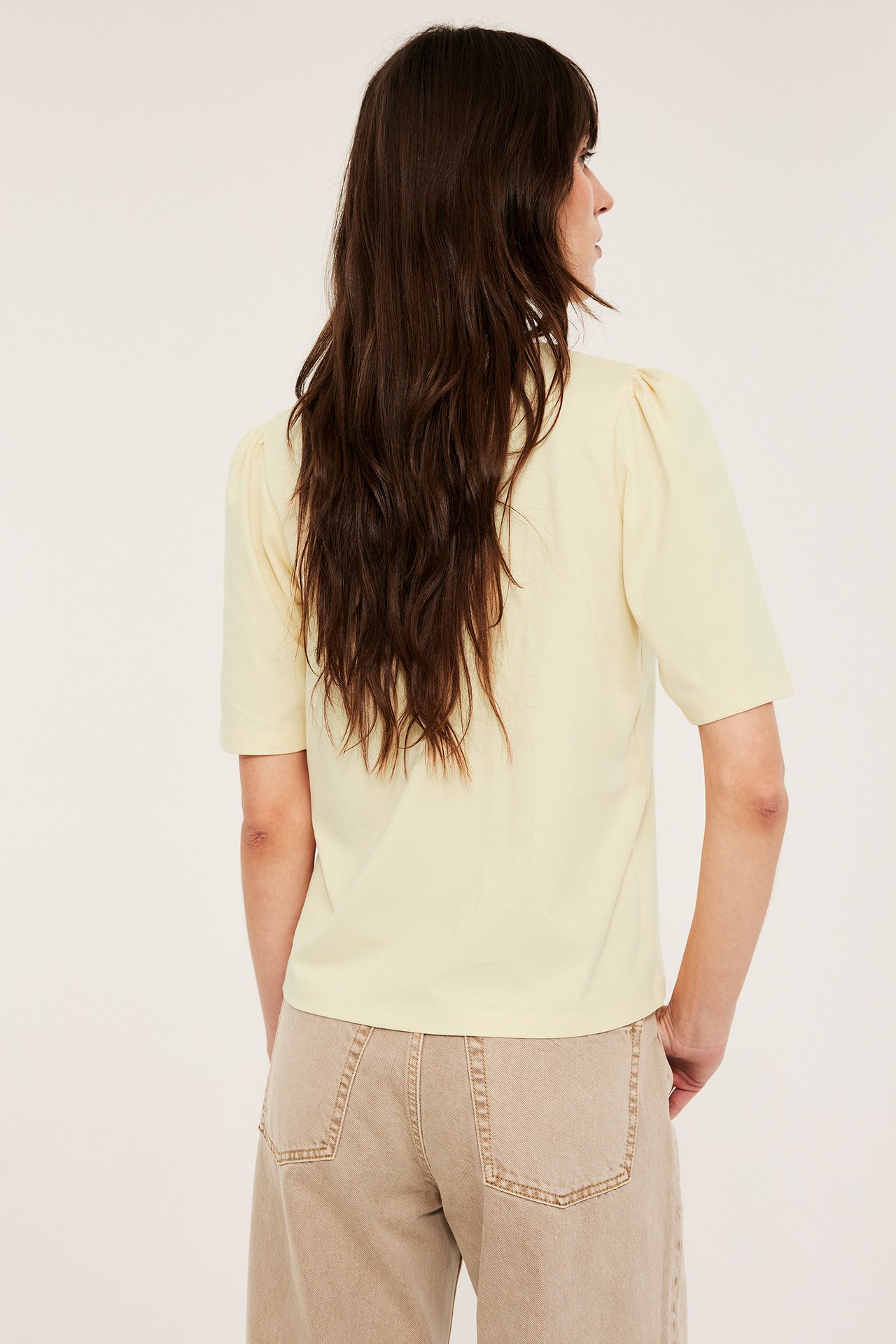 Ryggbilde av en kvinne i en lys gul bomullstopp med puffermer og beige vide bukser.