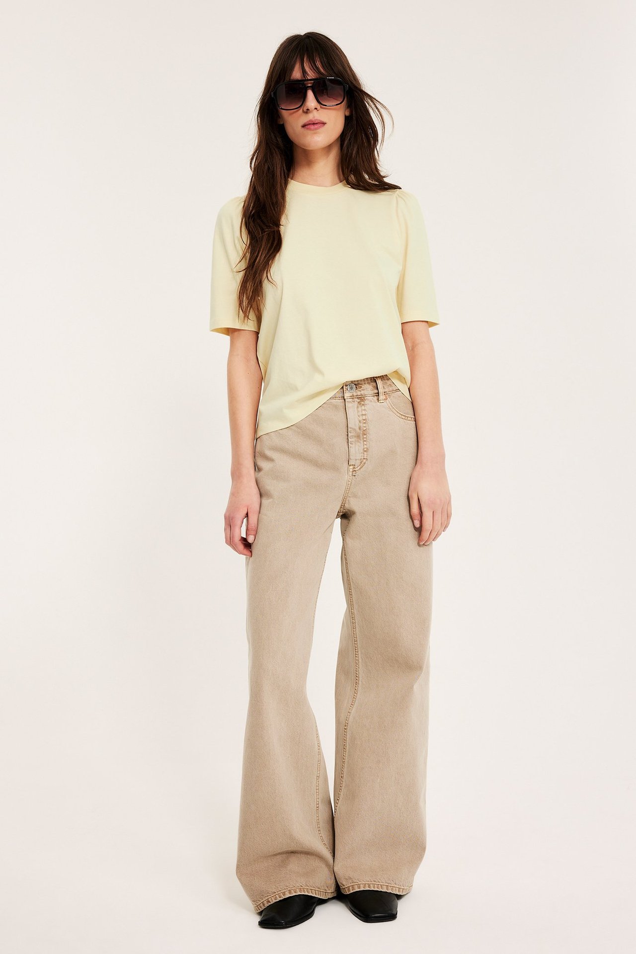 Frontalvisning: Kvinne i lys gul topp med puffermer, beige vide jeans, solbriller og sko.