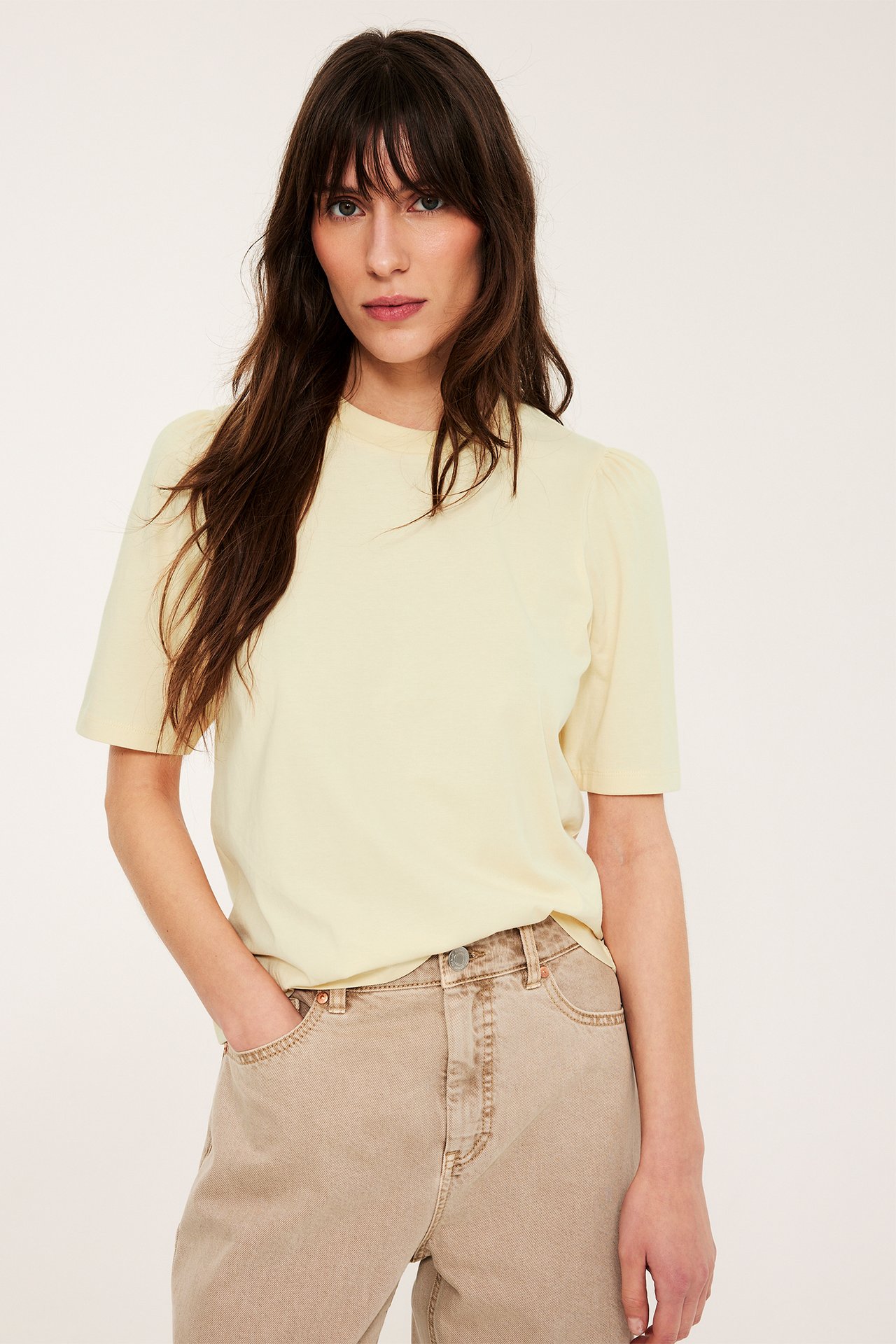 Frontbilde av en kvinne i en lys gul bomullstopp med puffermer og lysebrune jeans.