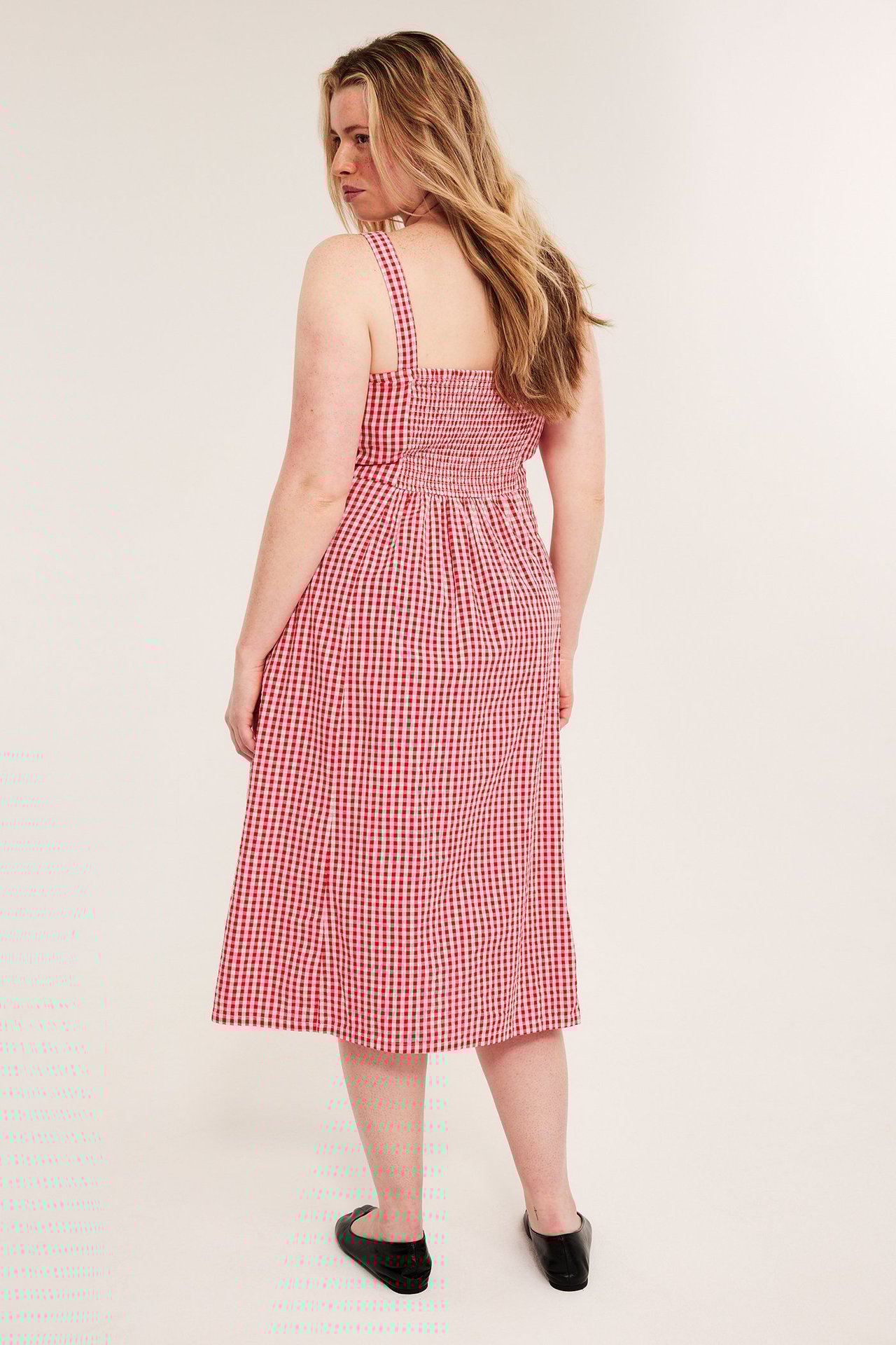 Ryggvy av kvinna i röd- och vitrutig bäckebölja midiklänning med smock i ryggen och svarta platta skor.