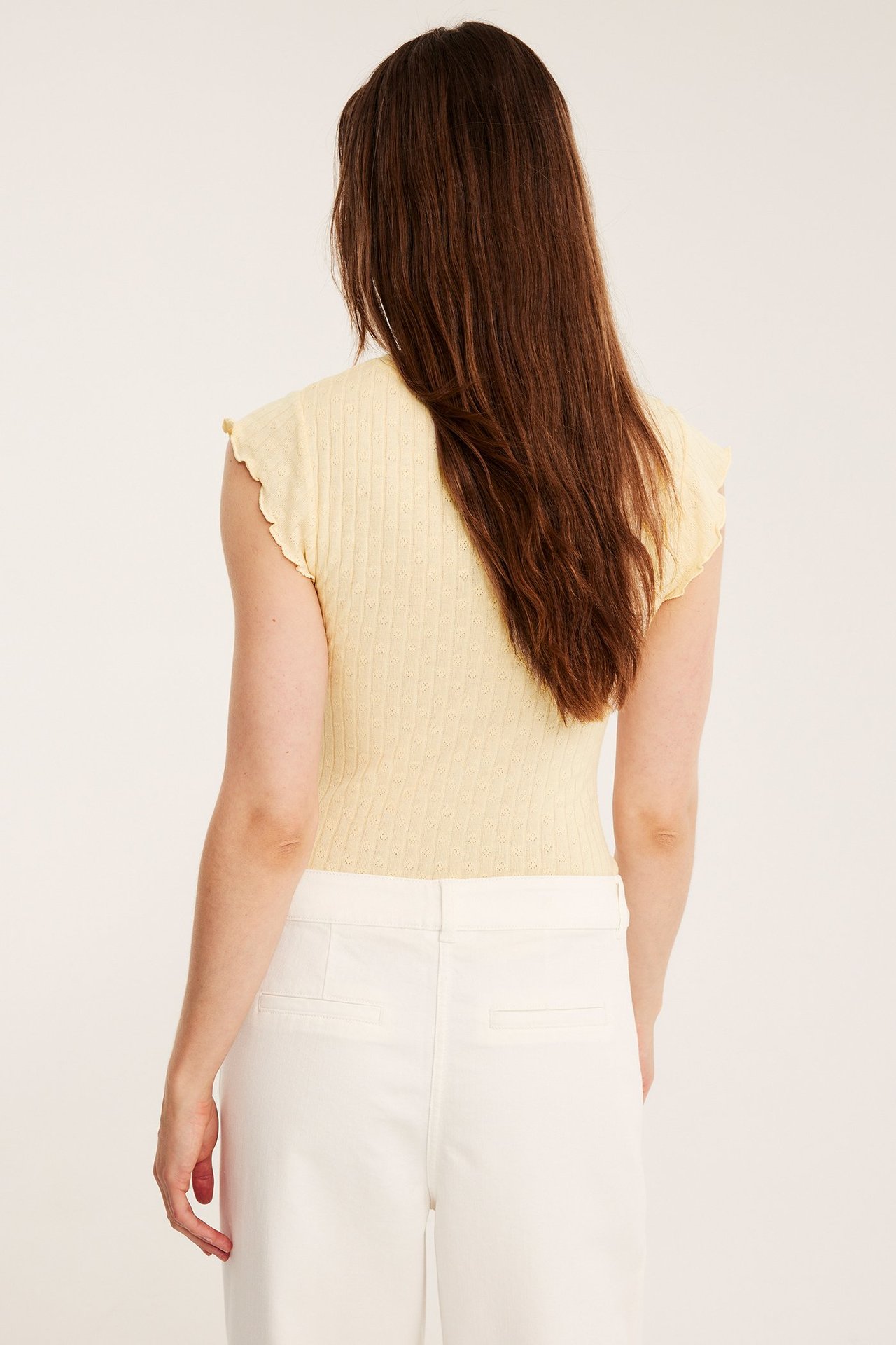 Naisen takakuva: vaaleankeltainen pointelle-toppi röyhelöhihoilla ja valkoiset housut.
