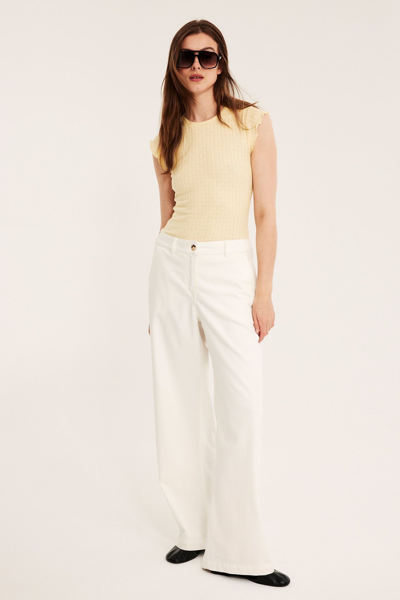 Nainen edestä: vaaleankeltainen pointelle-toppi, valkoiset leveälahkeiset housut ja aurinkolasit.