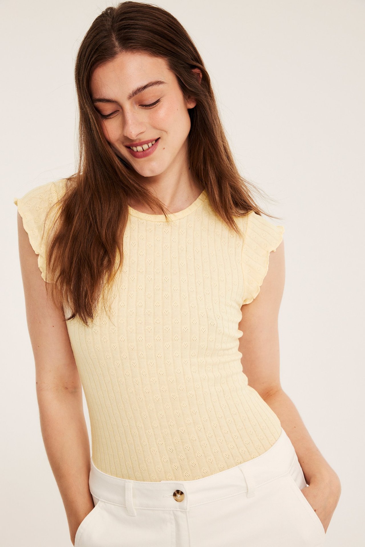 Framifrån, kvinna i ljusgul pointelle baby t-shirt med korta volangärmar och vita byxor.