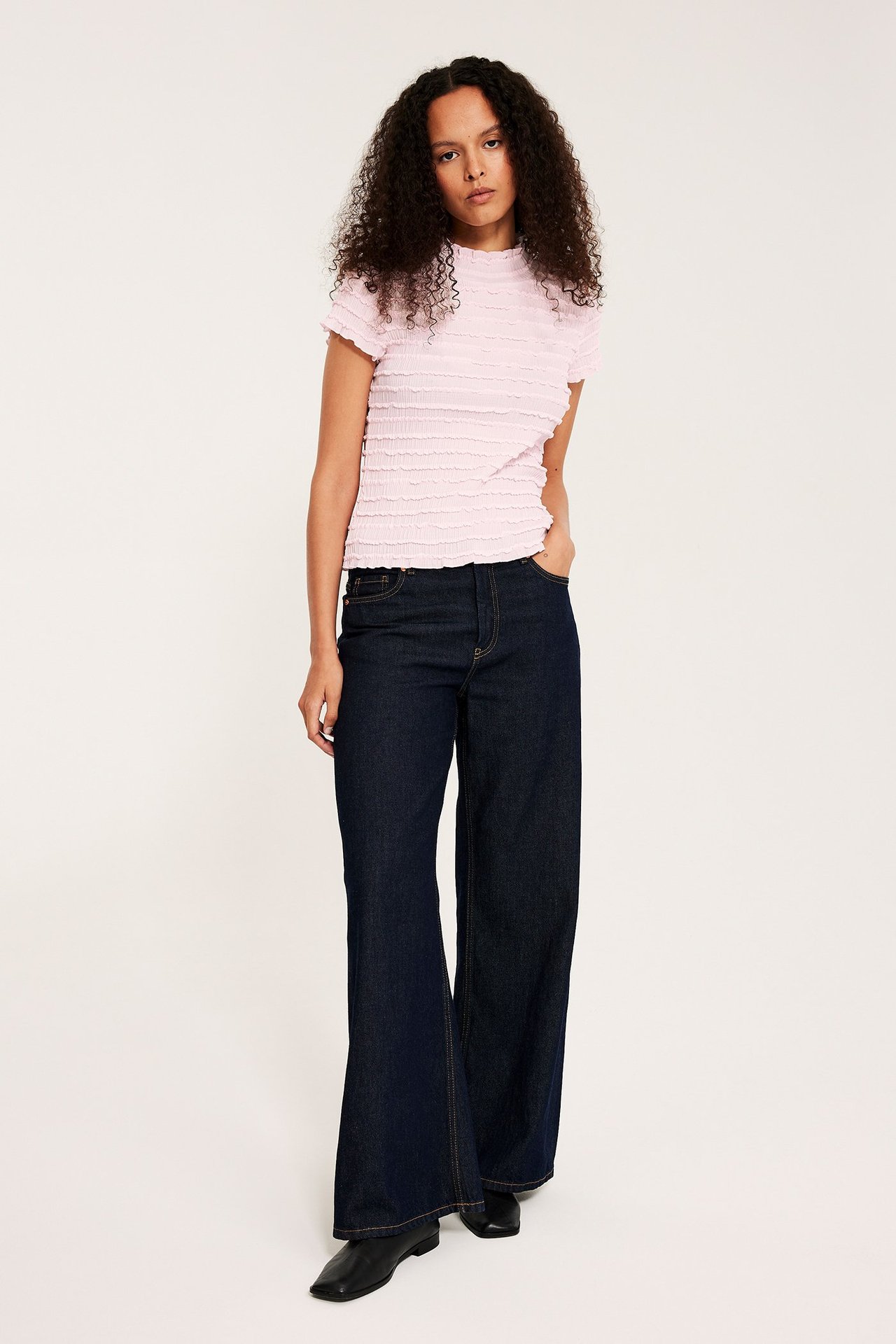Nainen edestä vaaleanpunaisessa rypytetyssä lyhythihaisessa topissa, tummansinisissä leveälahkeisissa farkuissa ja mustissa kengissä.