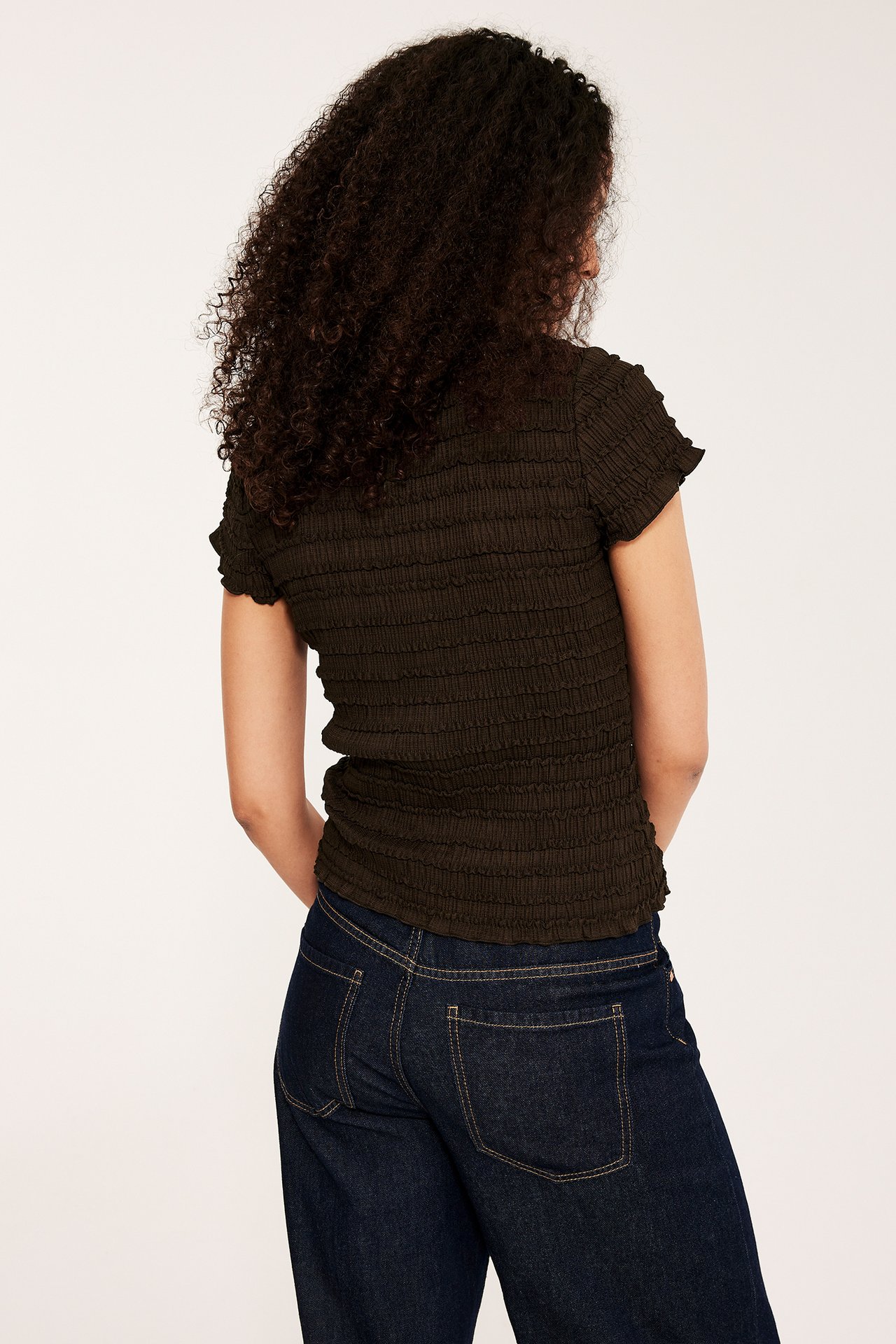 Nainen takaapäin, yllään tummanruskea rakenteellinen toppi ja tummansiniset farkut.