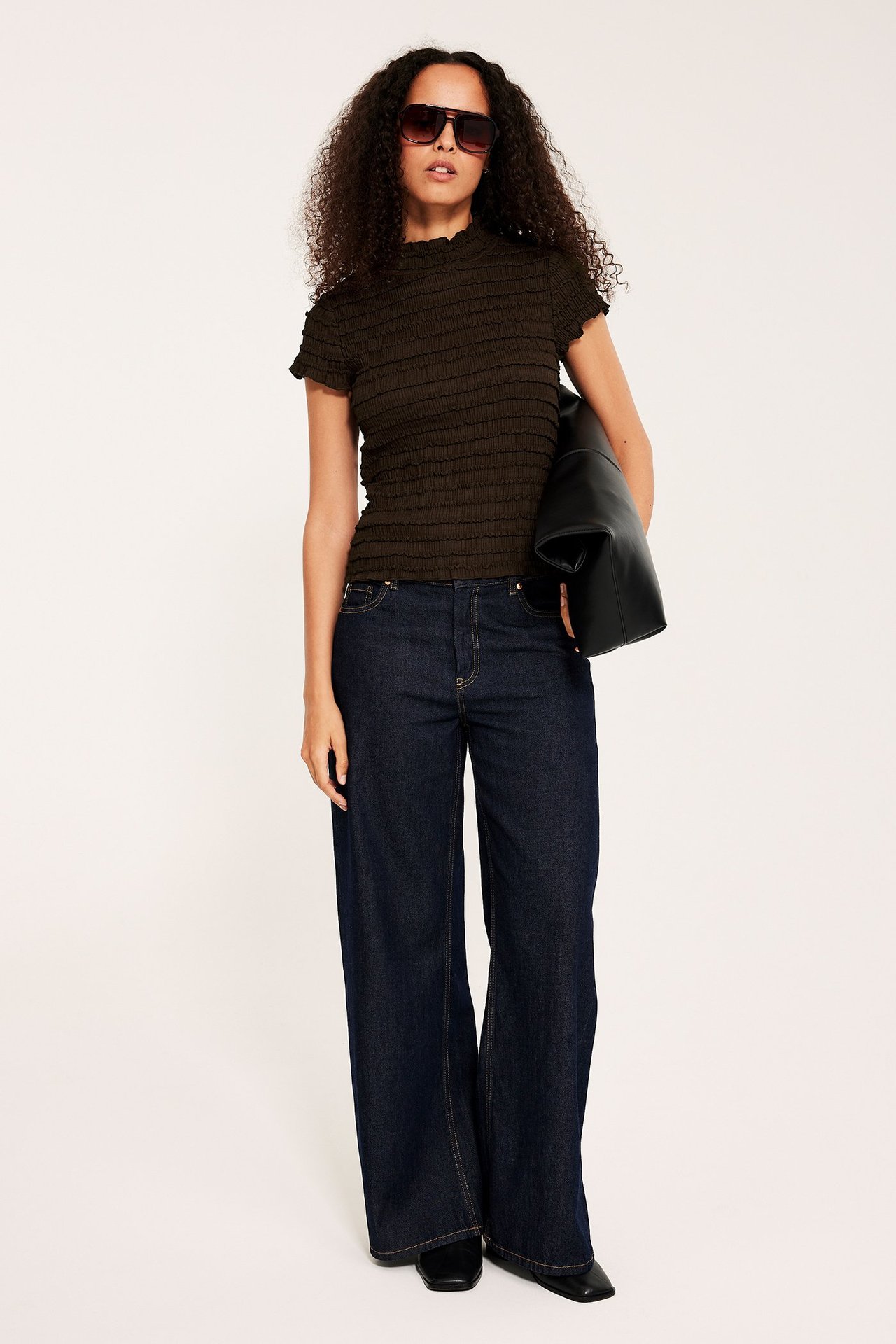 Nainen tummanruskeassa strukturoidussa topissa, tummansinisissä farkuissa, mustissa aurinkolaseissa, musta takki.