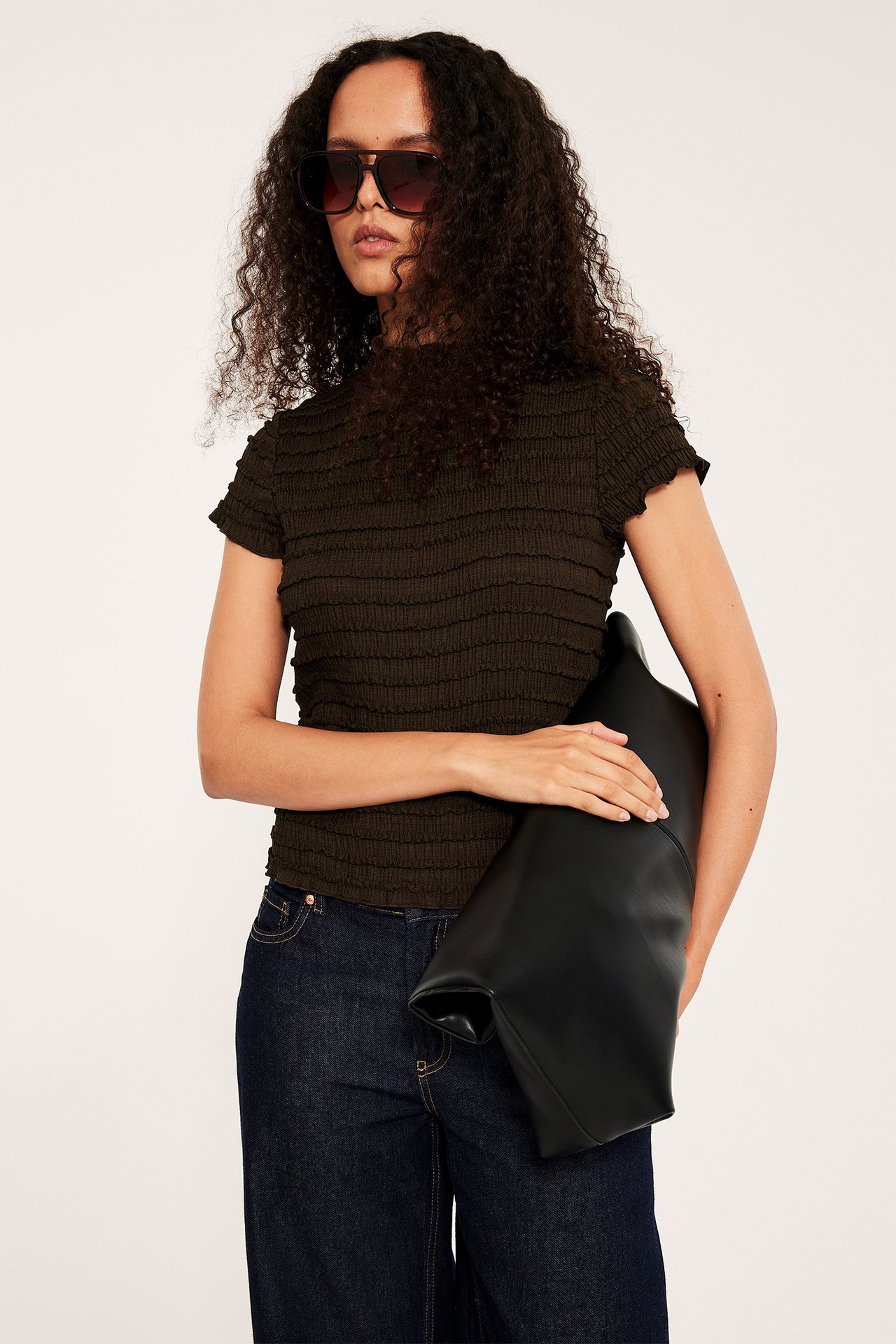 Nainen yllään tummanruskea kuvioitu toppi, tummat farkut, mustat aurinkolasit, pitää mustaa laukkua.