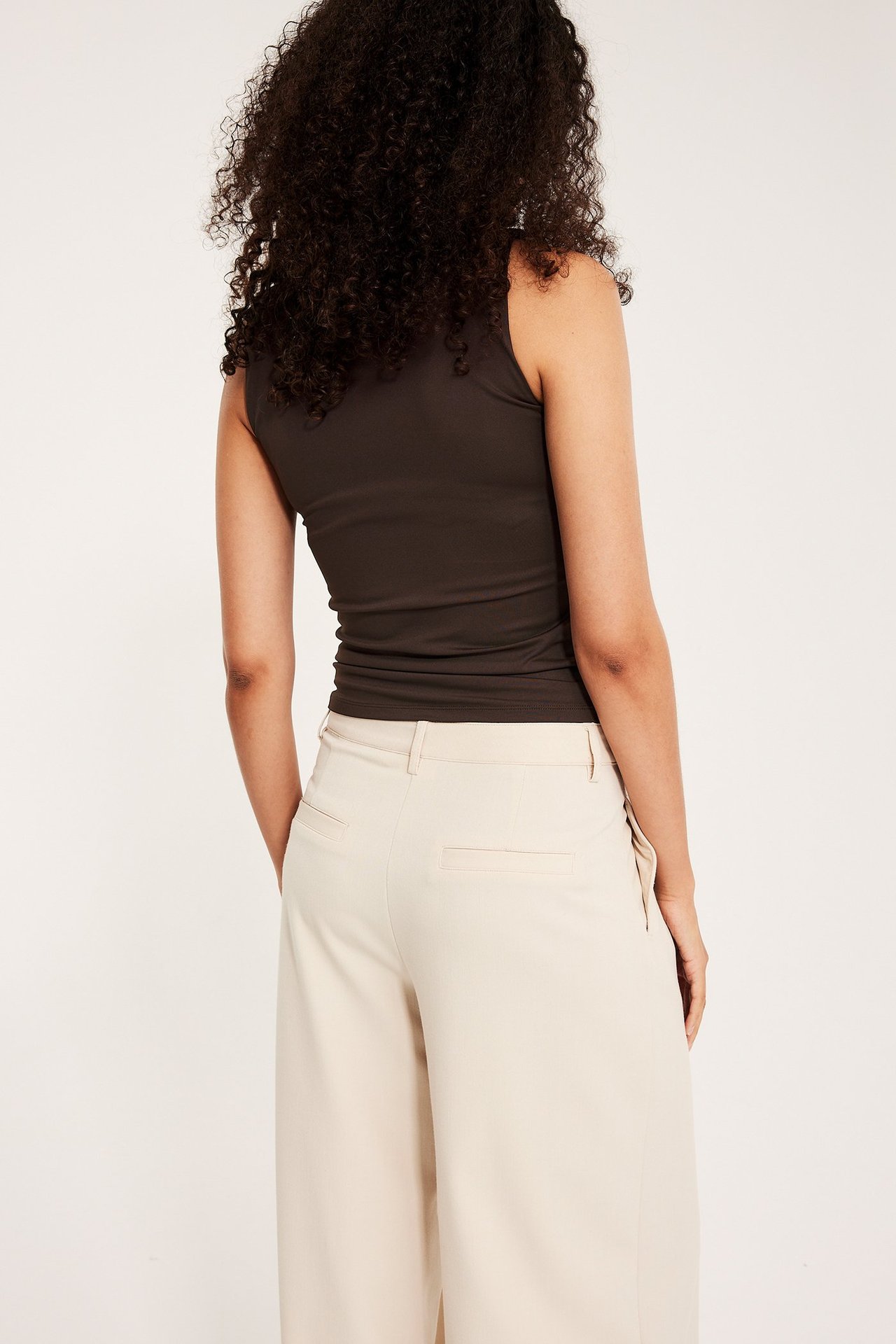 Nainen takaa, tiukka tummanruskea toppi ja vaaleanbeiget leveälahkeiset housut.