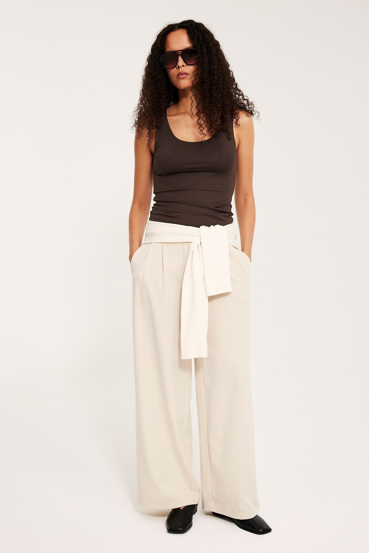 Nainen pukeutuneena tummanruskeaan tyköistuvaan toppiin, beigenvärisiin leveisiin housuihin ja solmittavaan vyöhön, mustiin kenkiin ja aurinkolaseihin.