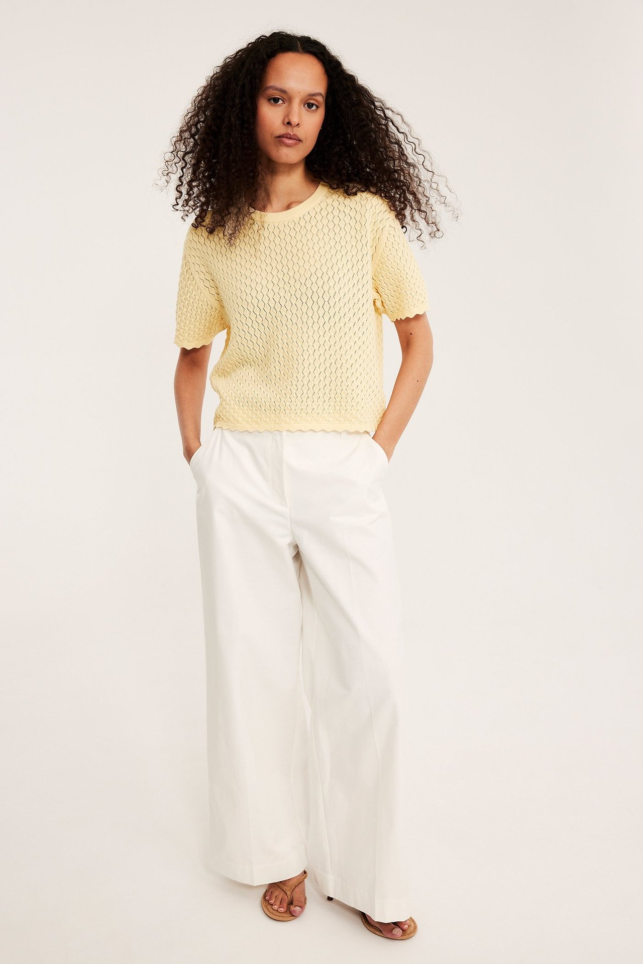 Widok z przodu kobiety w jasnożółtym dzianinowym swetrze z krótkim rękawem, białych spodniach i brązowych sandałach.