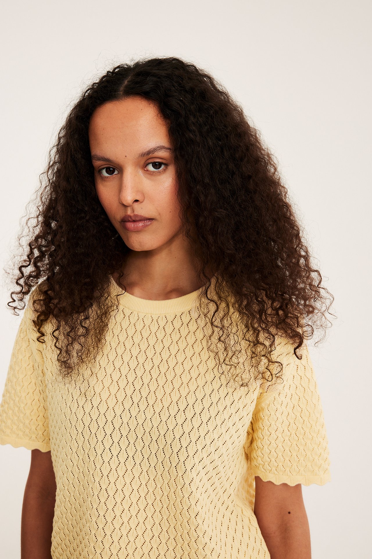 Lähikuva edestä naisesta yllään vaaleankeltainen kuviollinen lyhythihainen neulepusero, jossa on aaltoilevat reunat.