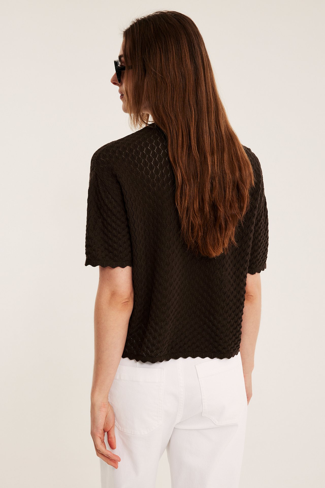 Rygg/sida vy av kvinna i mörkbrun mönsterstickad kortärmad tröja, vita byxor och svarta solglasögon.