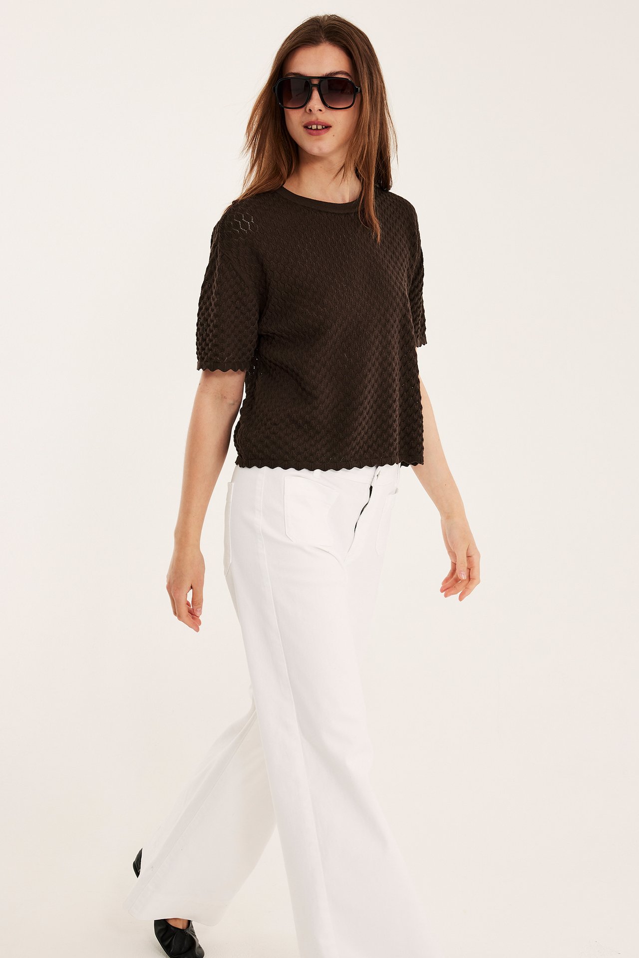 Edestä: nainen yllään tummanruskea kuviollinen lyhythihainen puuvillaneule, valkoiset leveälahkeiset housut ja mustat aurinkolasit.